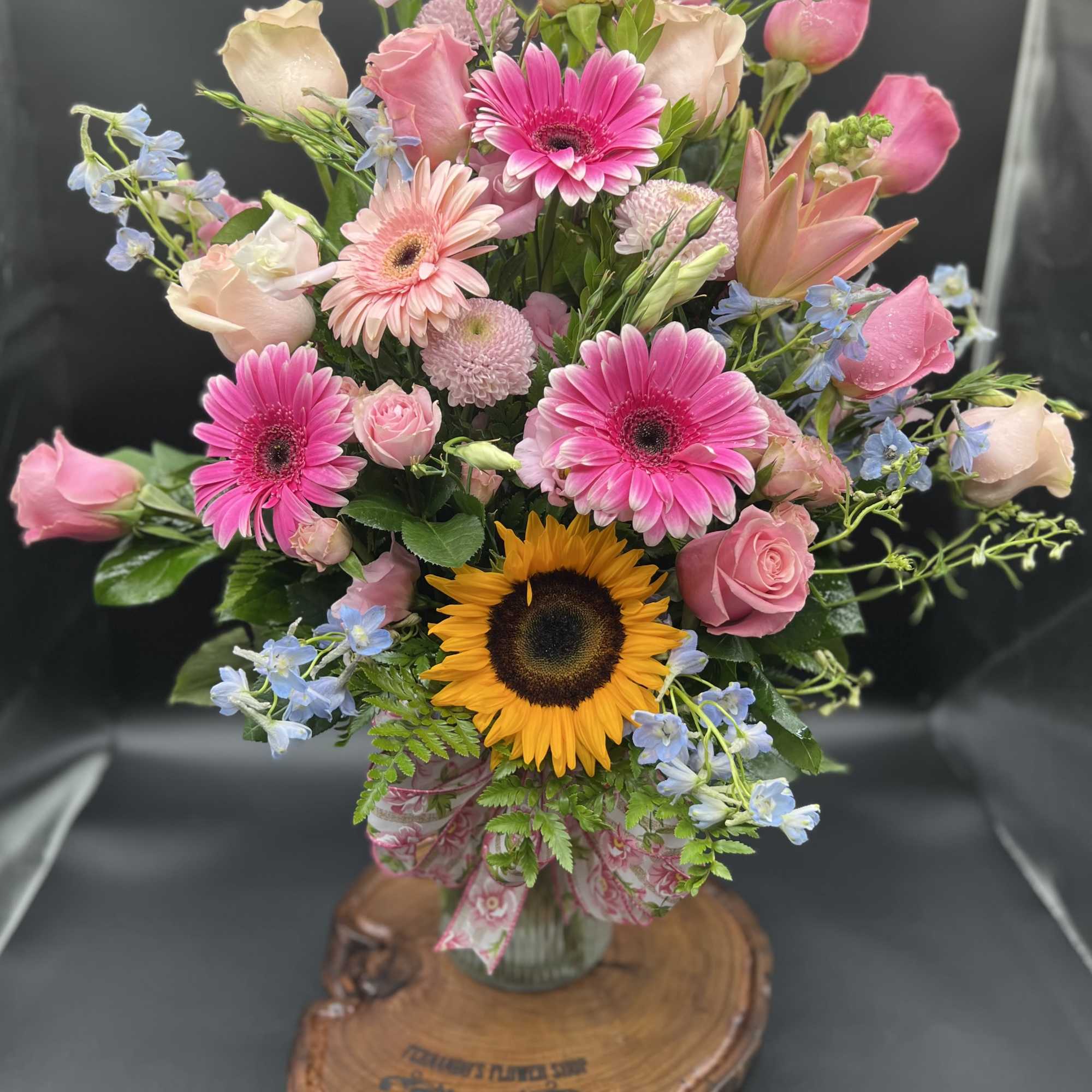 Tall mixed arrangement of pink roses, gerbera daisies, and a sunflower in a glass vase with ribbon