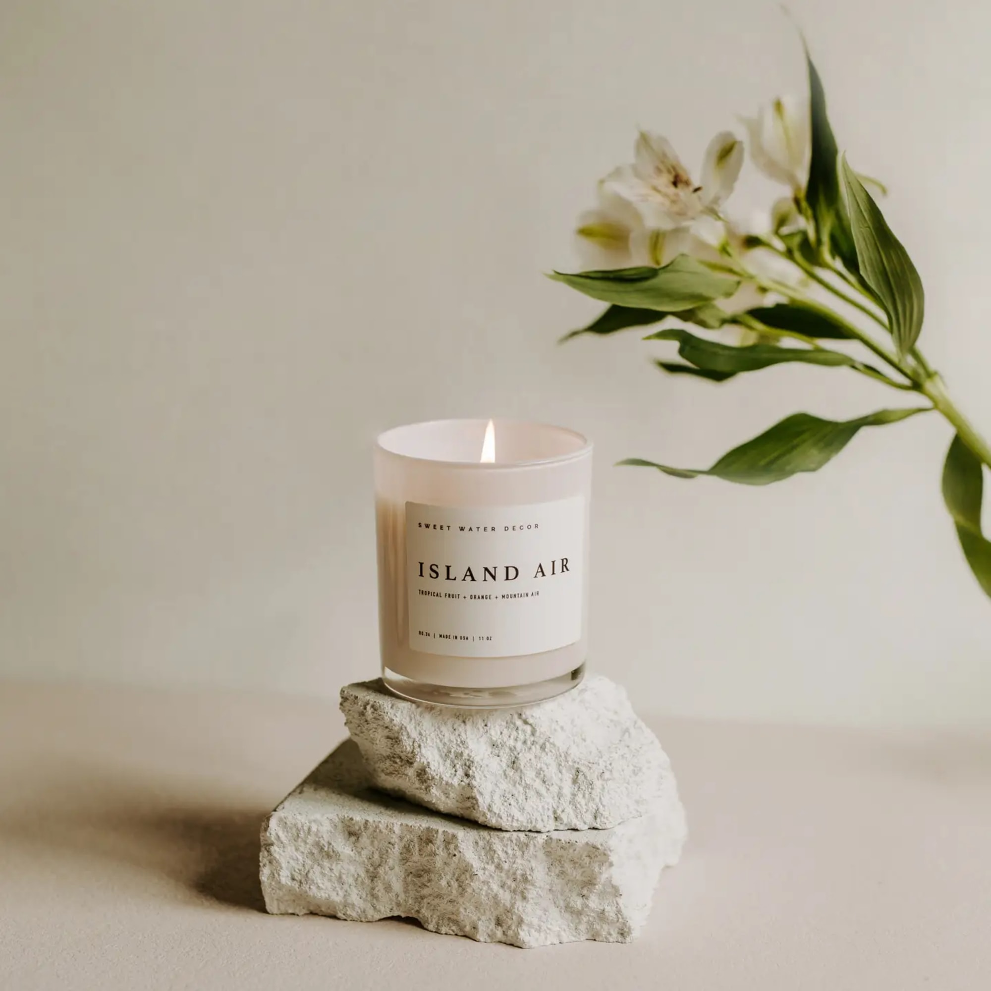 Lit candle in a glass jar beside a white flower stem on stacked stones
