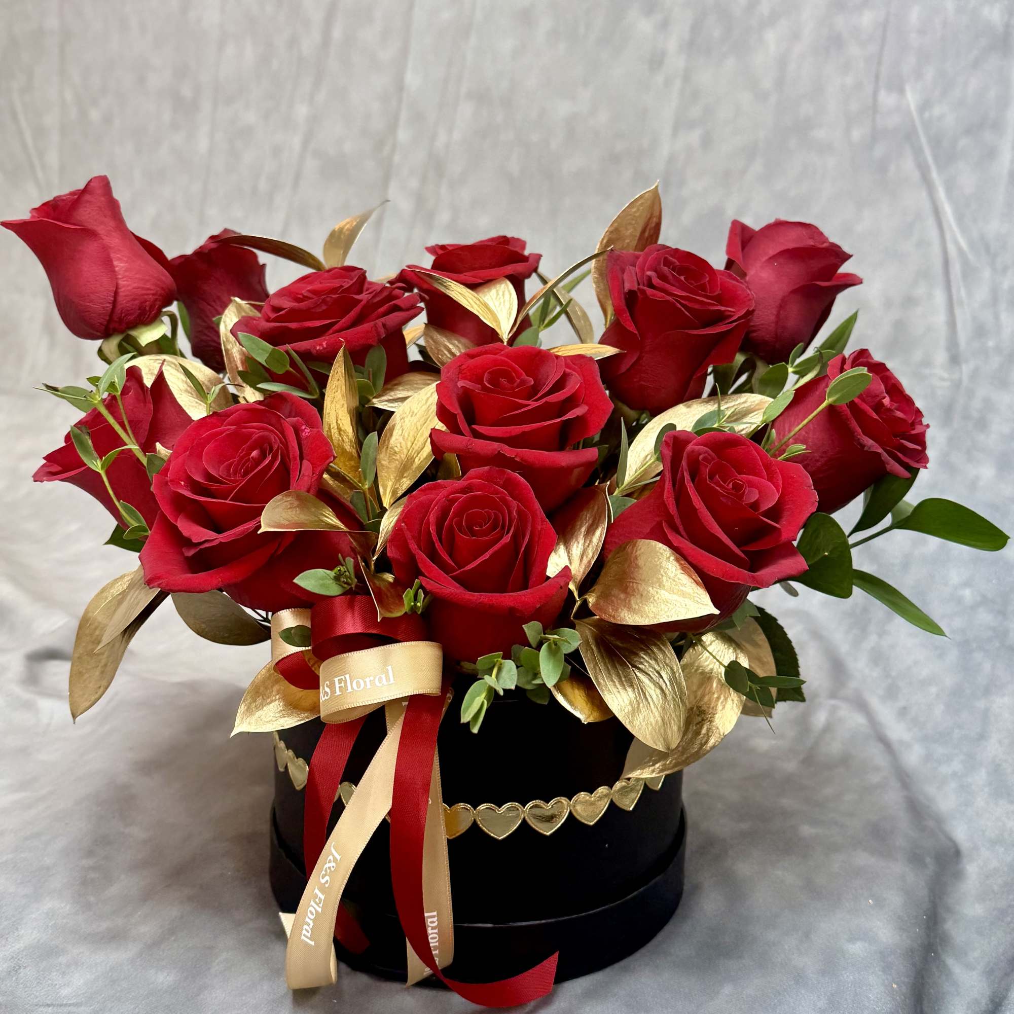 Red roses arranged in a black hat box with gold leaves and ribbon