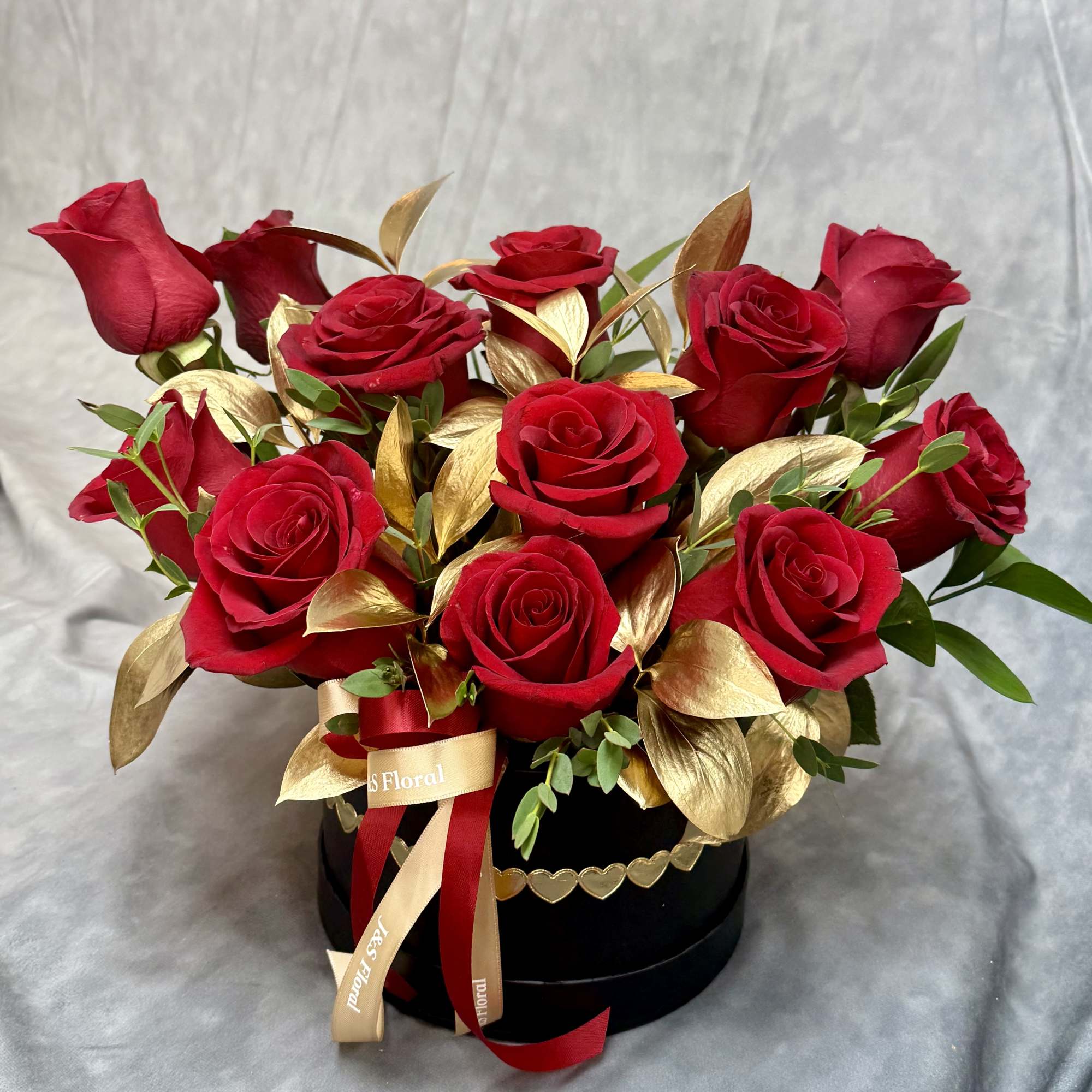 Red roses arranged in a black round box with gold leaves and ribbon