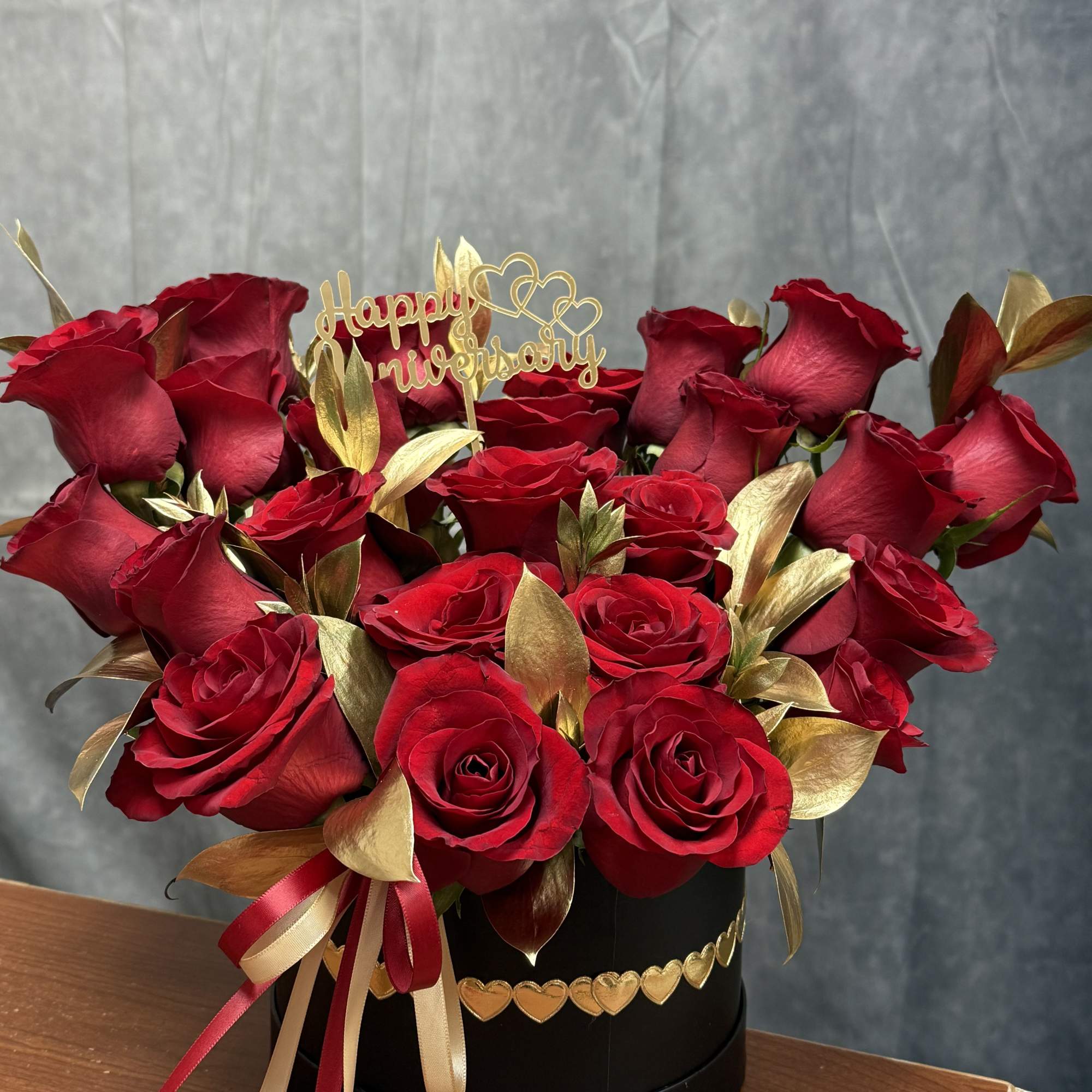 Red roses arranged in a black hat box with gold accents and a Happy Anniversary topper