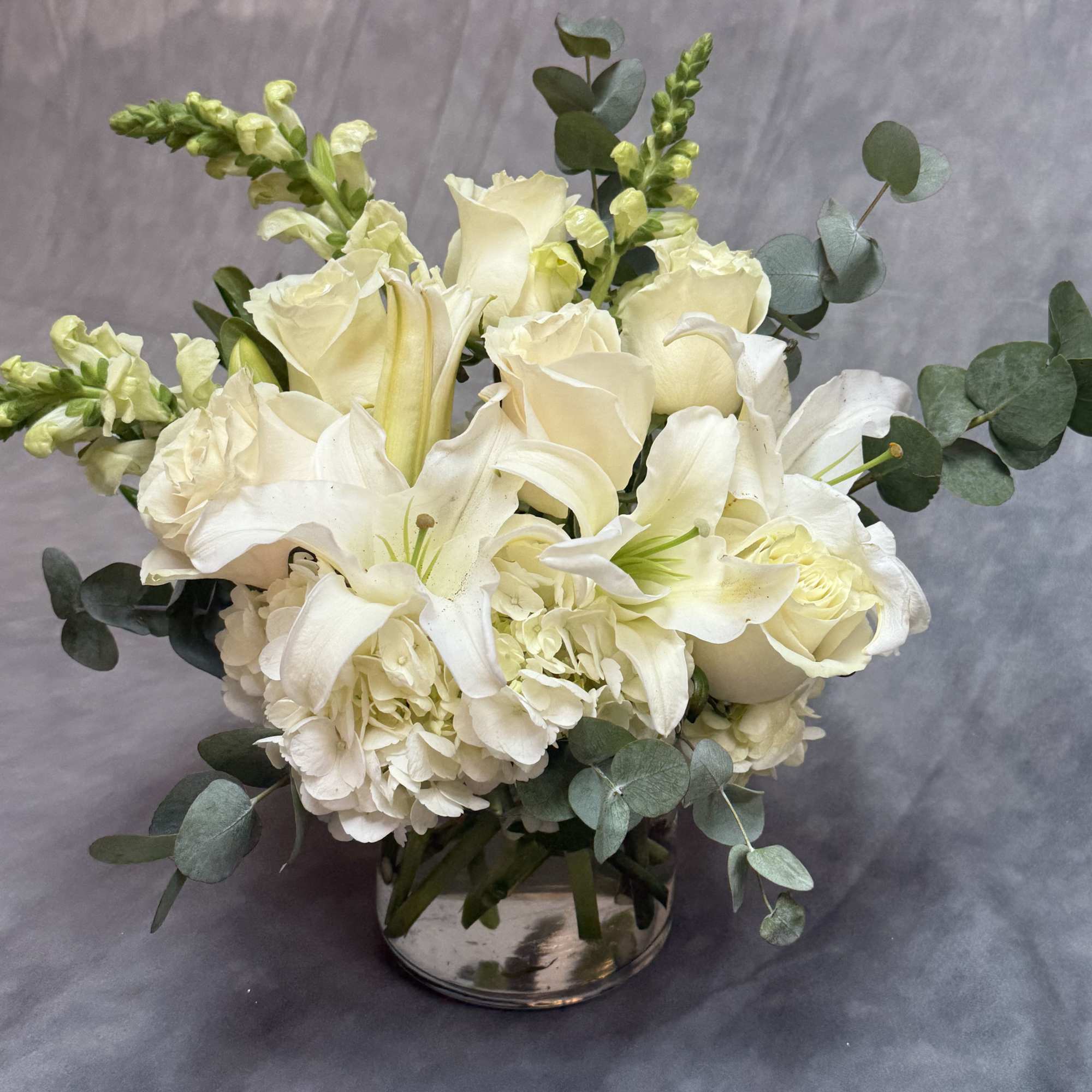 Low white arrangement of roses, lilies, hydrangeas, and snapdragons in a clear glass cylinder vase