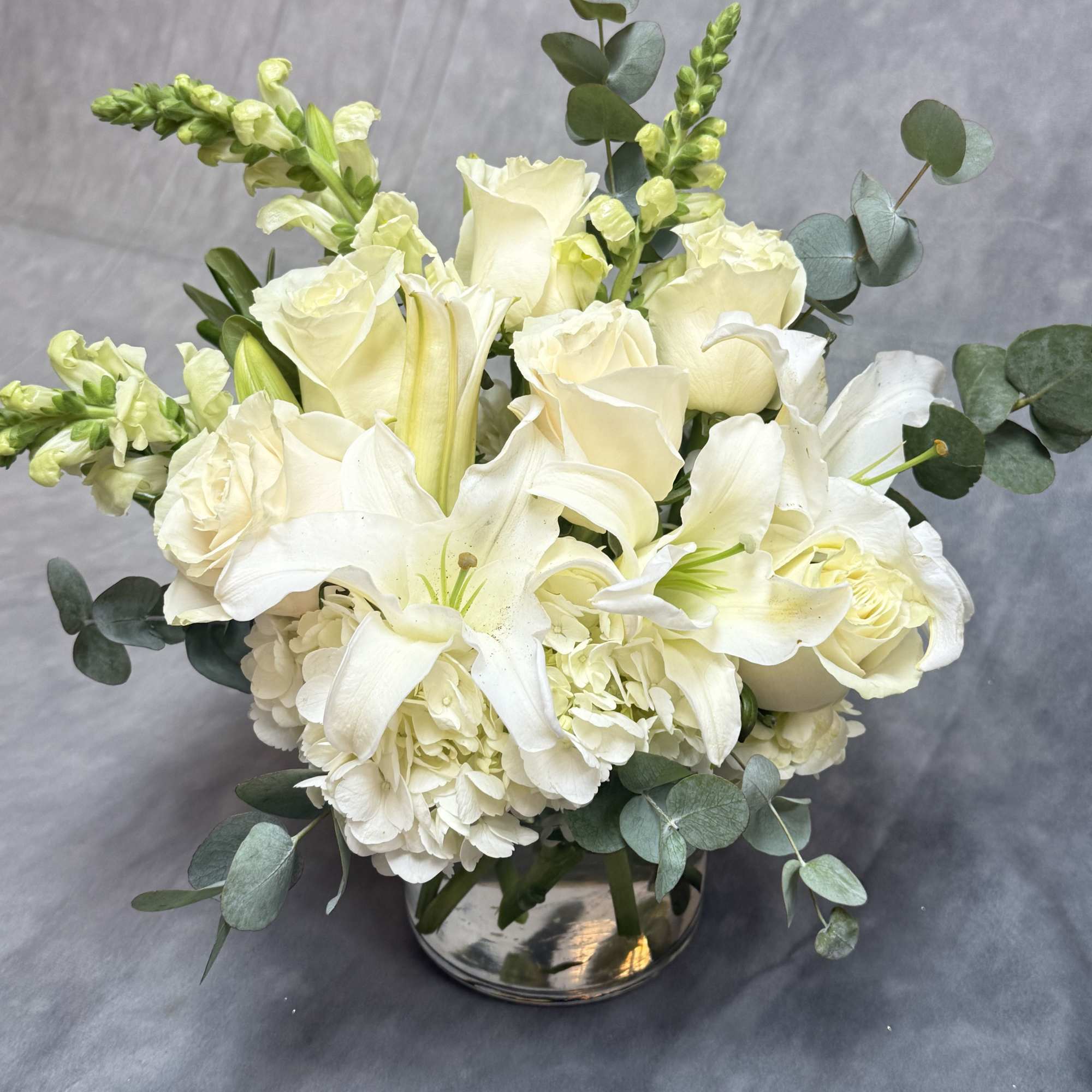 Low arrangement of white lilies, roses, hydrangeas, and snapdragons in a silver bowl vase