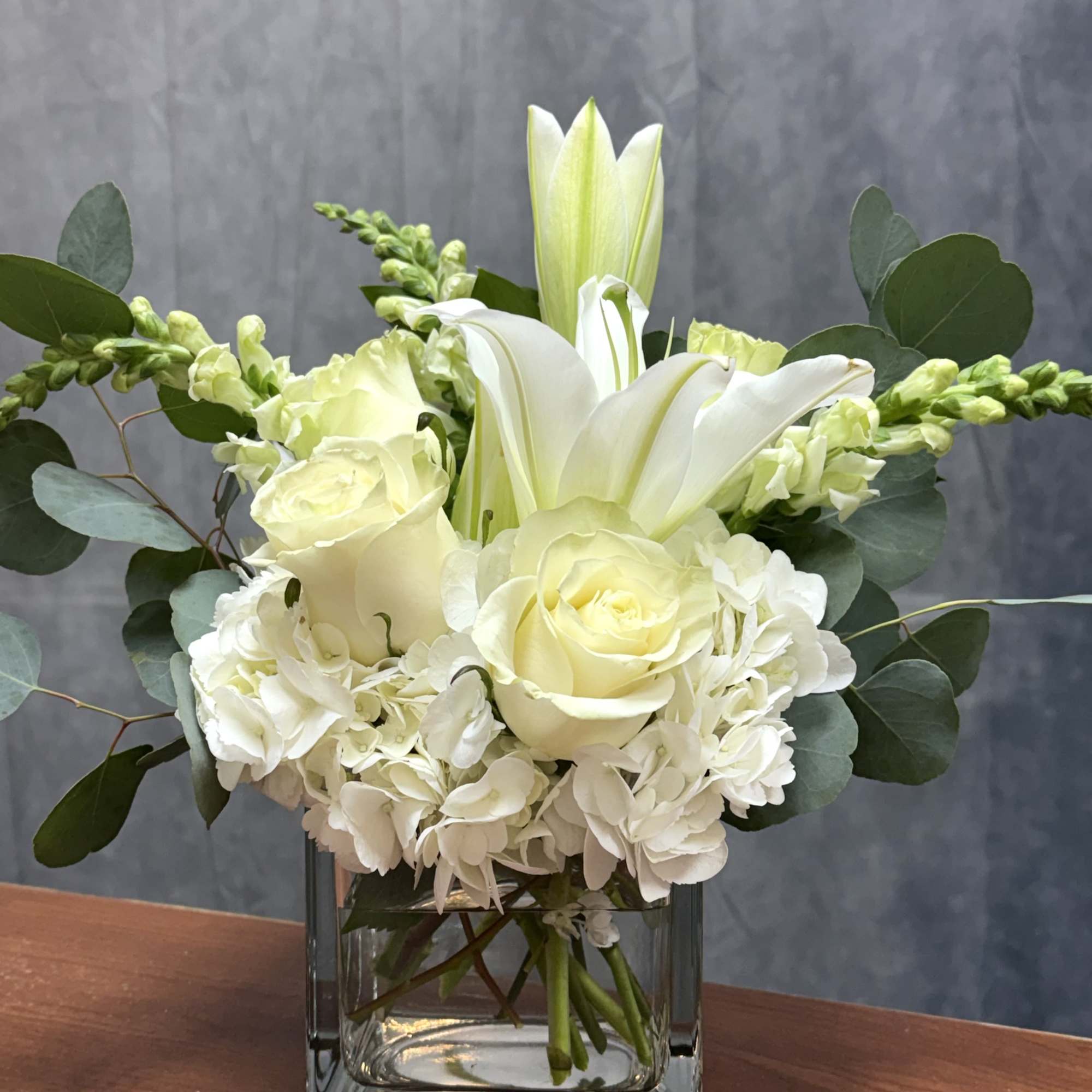 White lilies, roses, and hydrangeas arranged in a clear glass cube vase