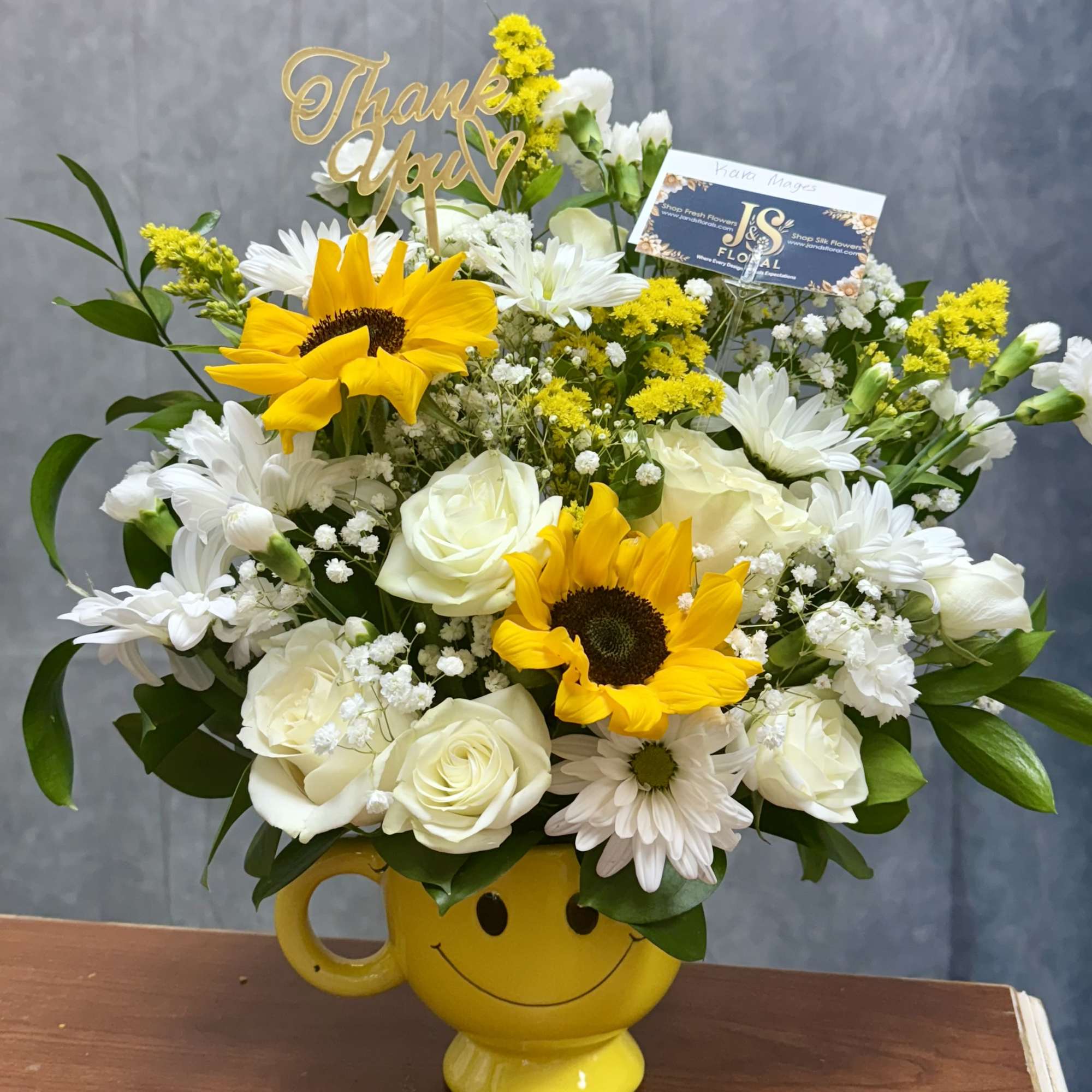 Cheerful bouquet of sunflowers, white roses, and daisies in a yellow smiley mug with a thank you pick.