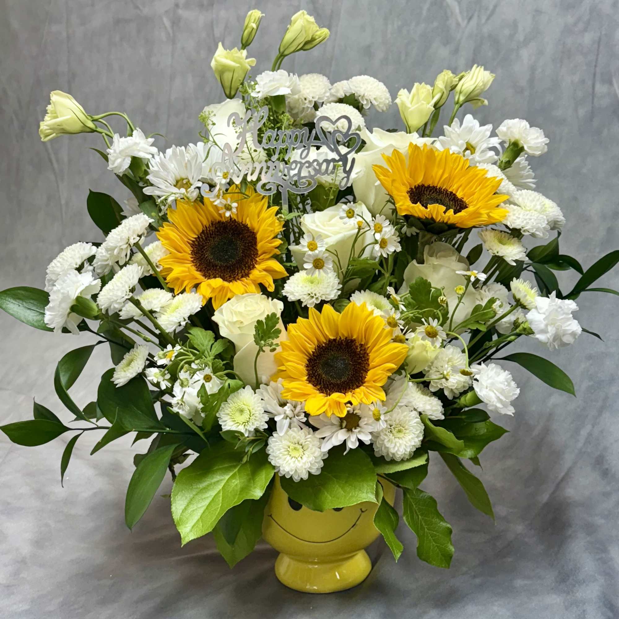 Yellow and white sunflower and rose arrangement in a yellow smiley mug with Happy Anniversary pick
