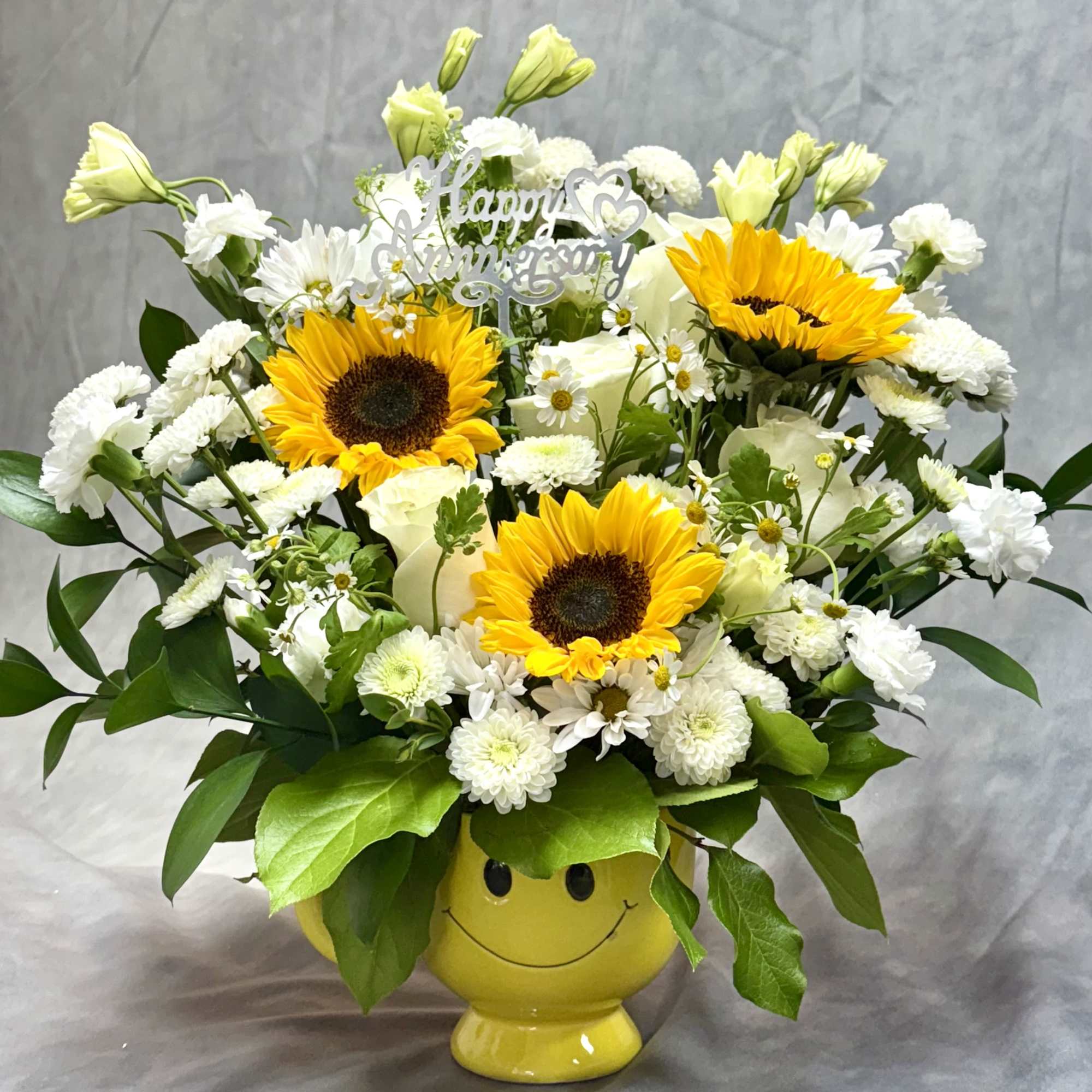 Cheerful yellow and white flower arrangement with sunflowers in a yellow smiley-face mug and Happy Anniversary pick.