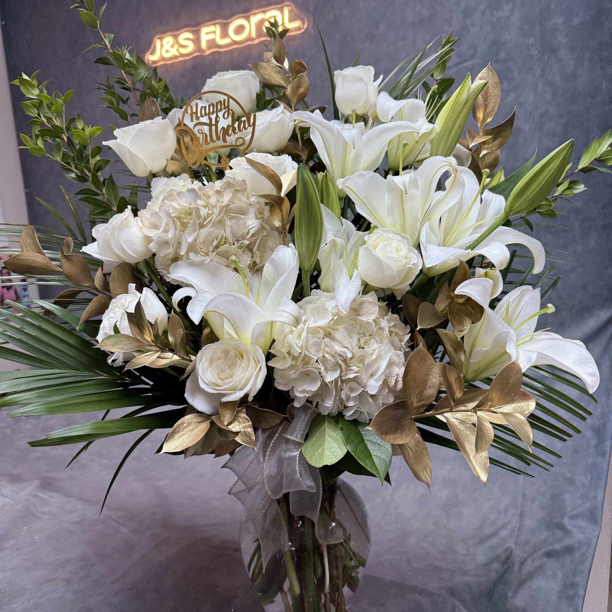 White lilies and roses in a glass vase with gold foliage and a Happy Birthday topper