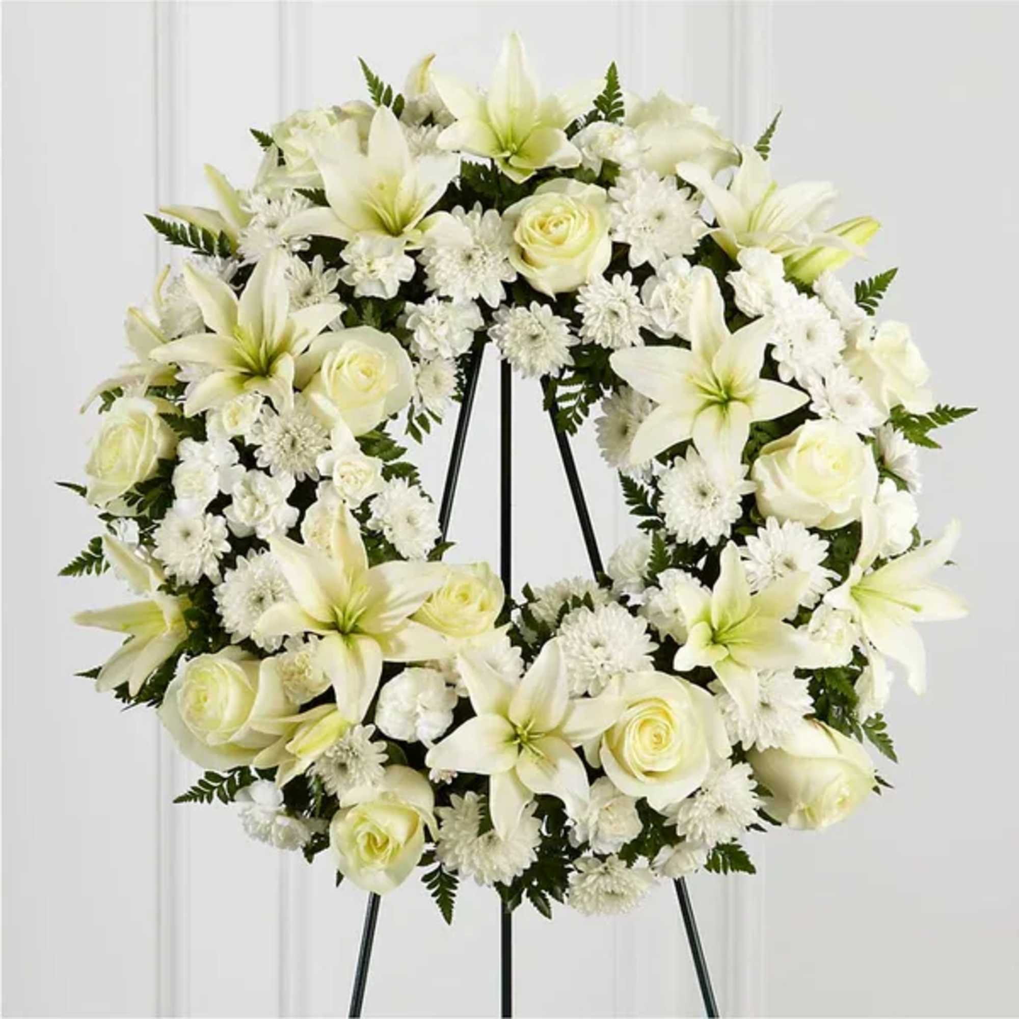 White floral wreath on a black stand with lilies and roses