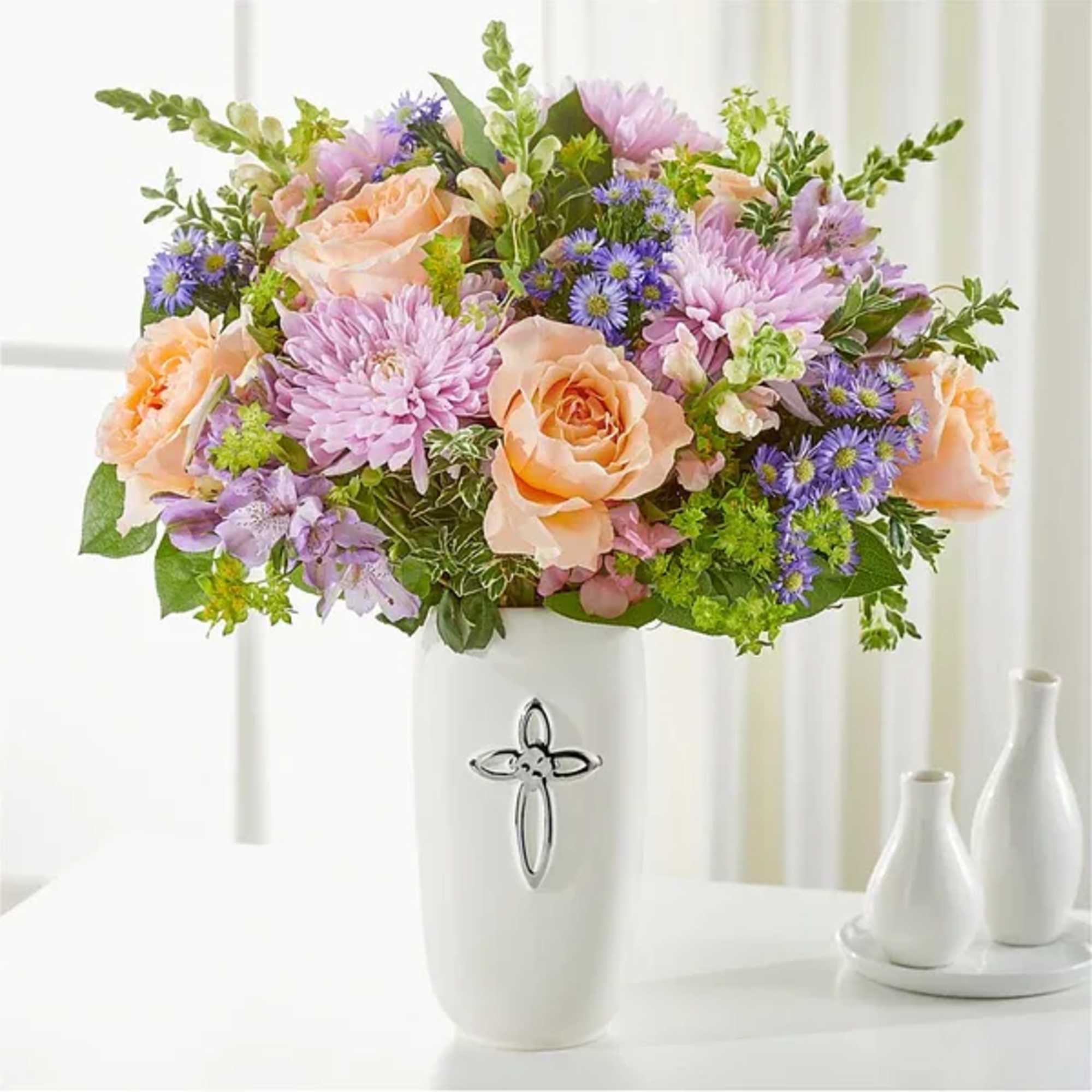 Pastel bouquet of roses and purple daisies in a white vase with a cross emblem