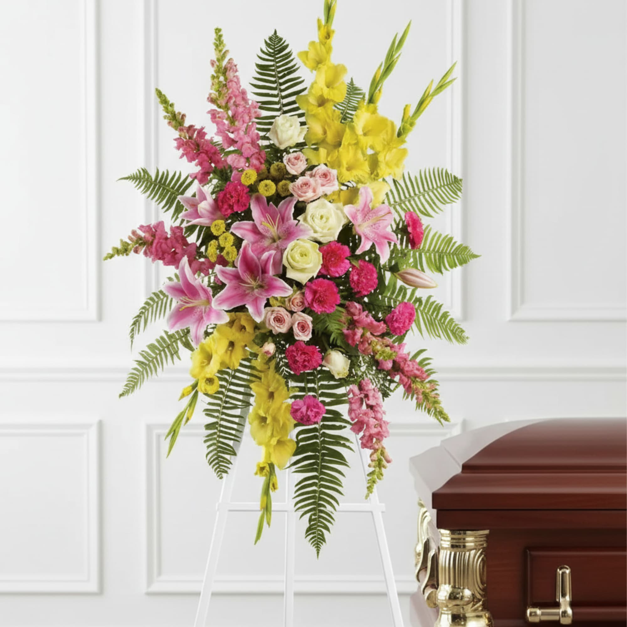 Standing spray of pink lilies, roses, and yellow flowers on a white easel beside a wooden casket.