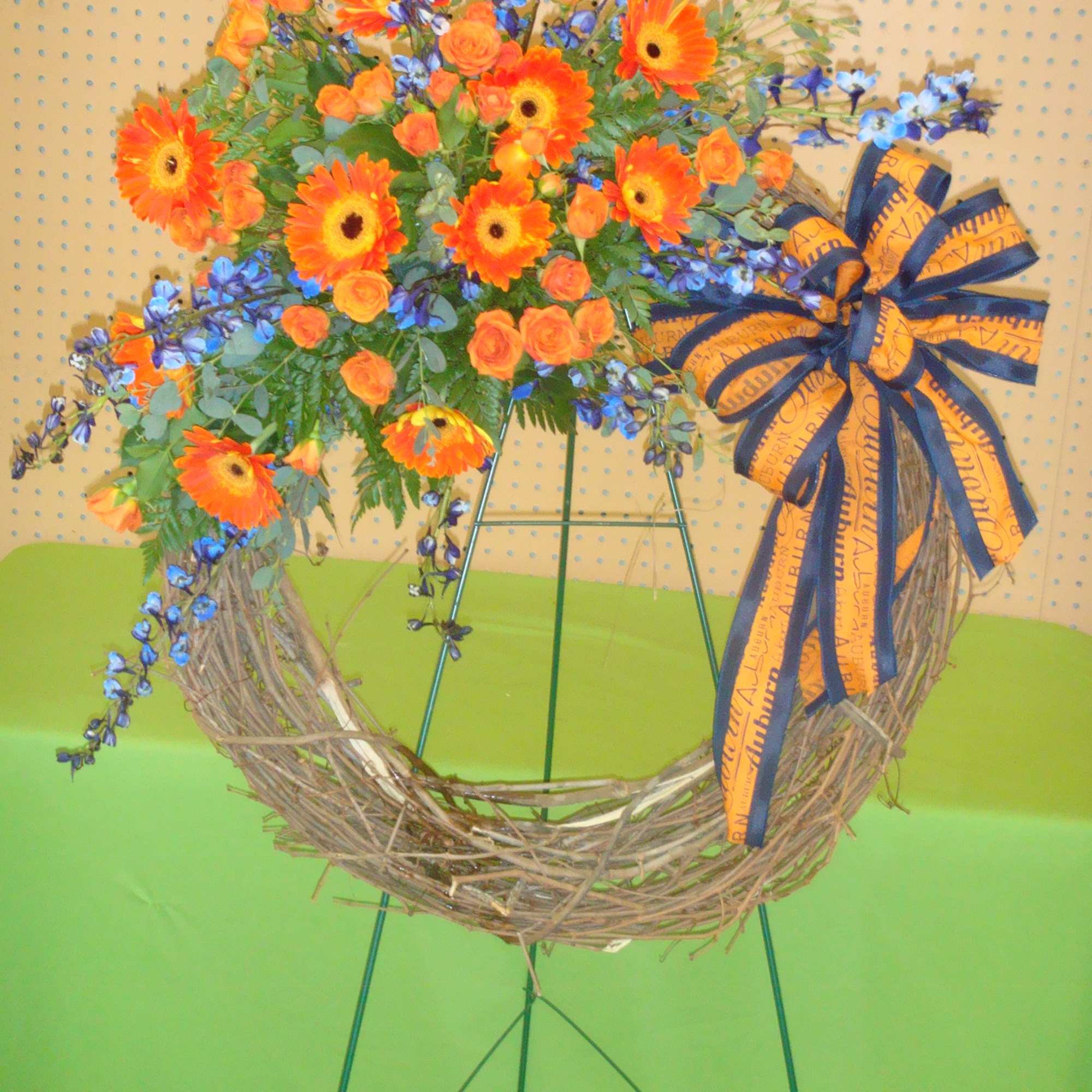 Grapevine wreath on an easel decorated with orange gerbera daisies, orange roses, blue flowers, and a large striped bow.