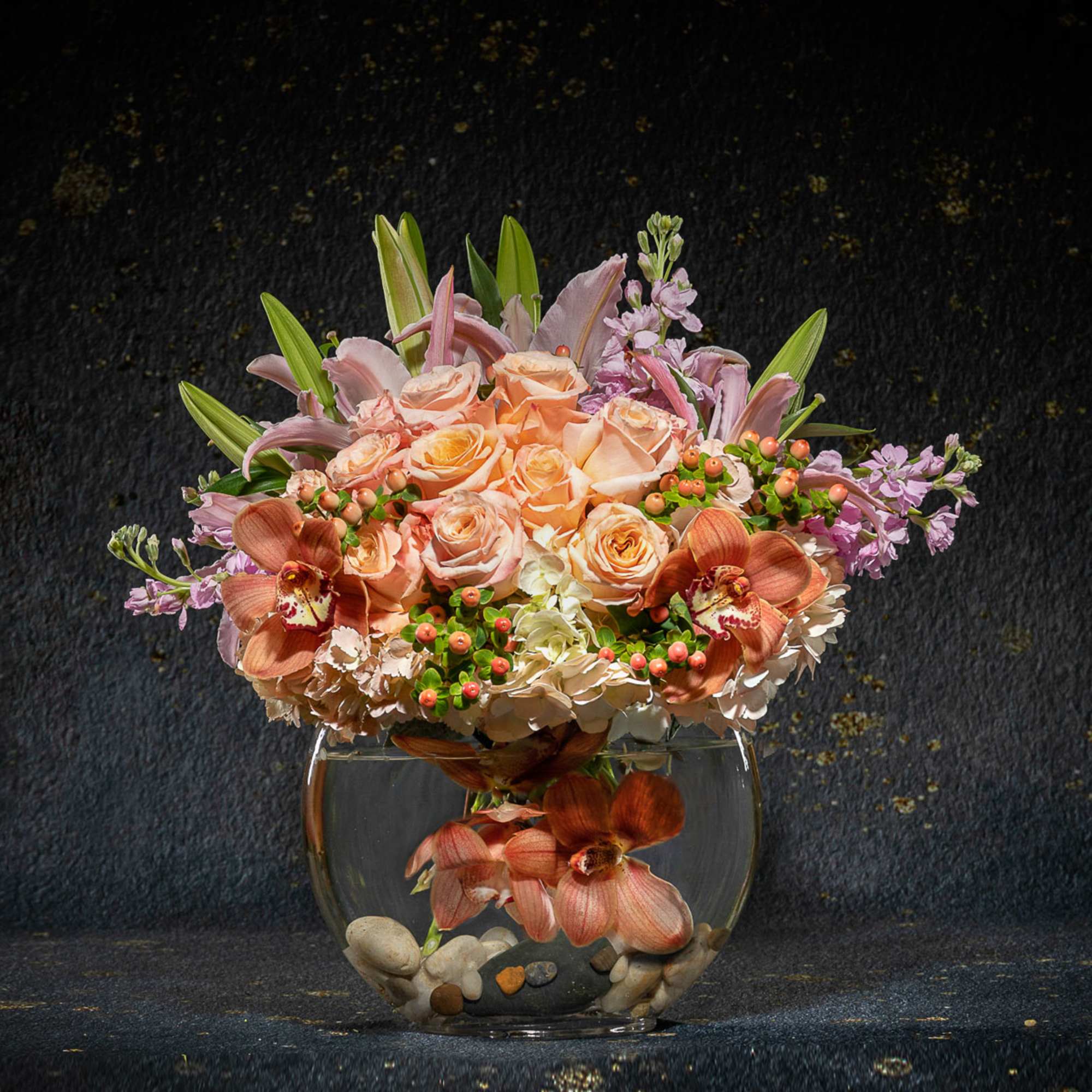 Peach roses and orchids arranged in a round glass vase with stones