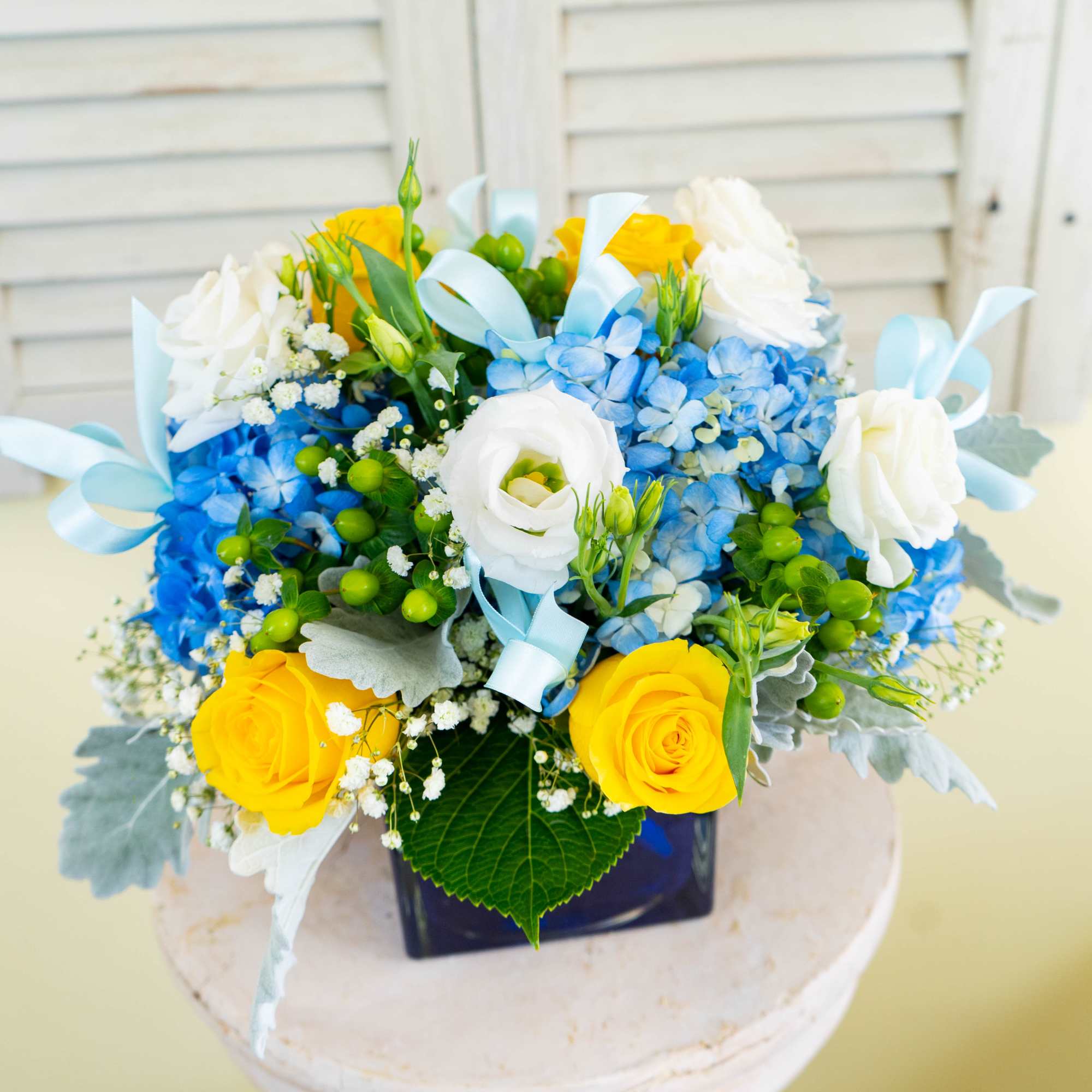 Blue and yellow floral arrangement in a dark square vase with ribbon accents