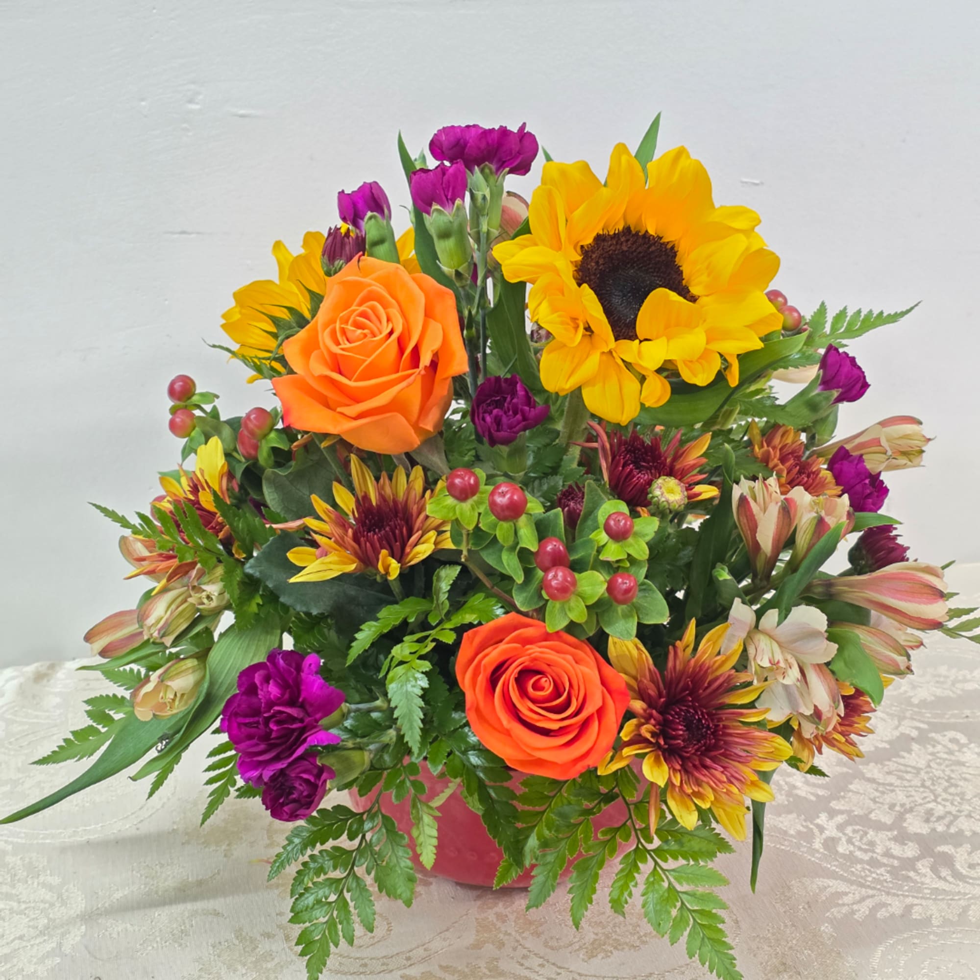 Mixed bouquet with sunflowers, orange roses, and purple blooms in a pink container