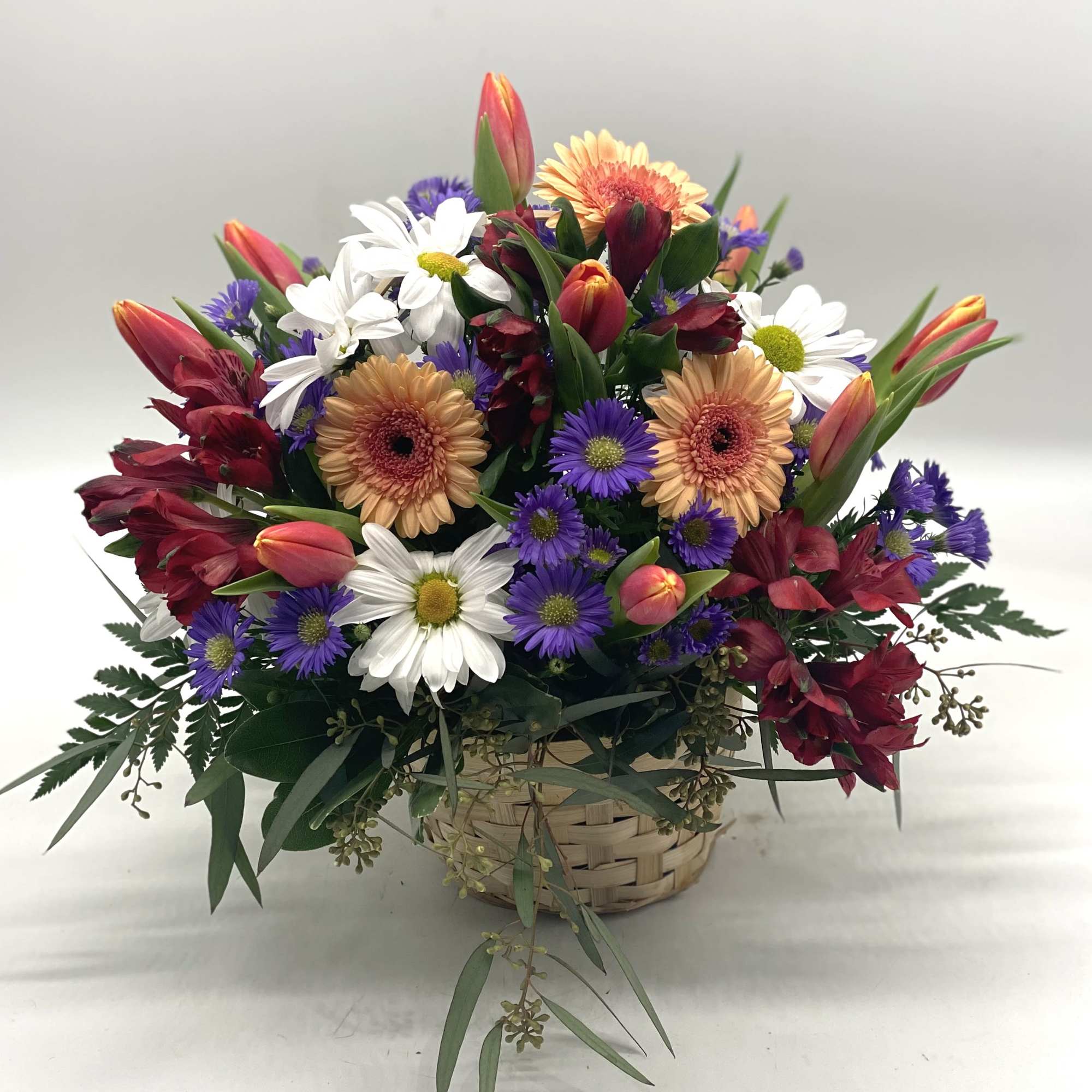 Mixed bouquet in a woven basket with daisies, gerberas, tulips, and purple asters