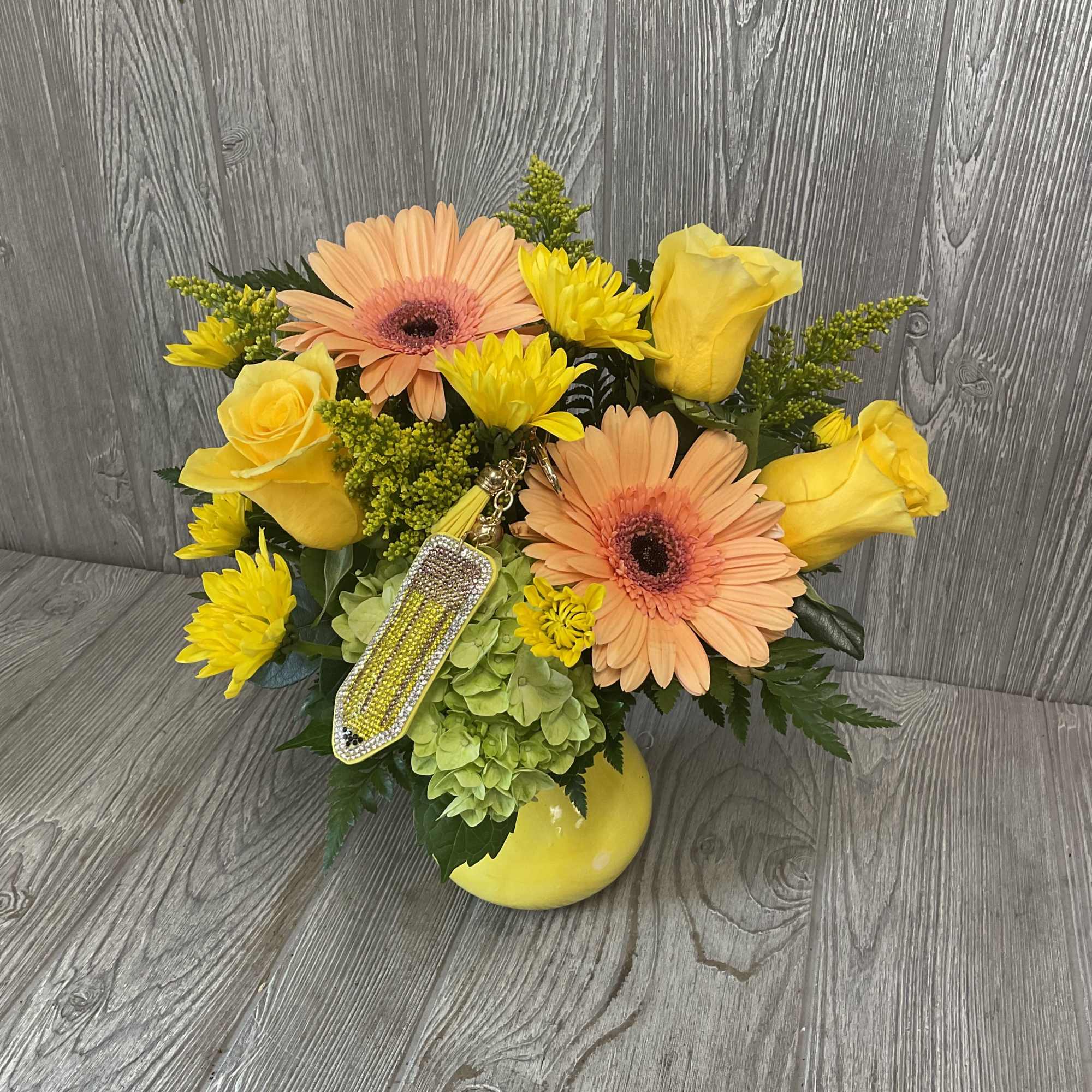 Yellow roses and peach gerbera daisies in a low arrangement with a decorative charm