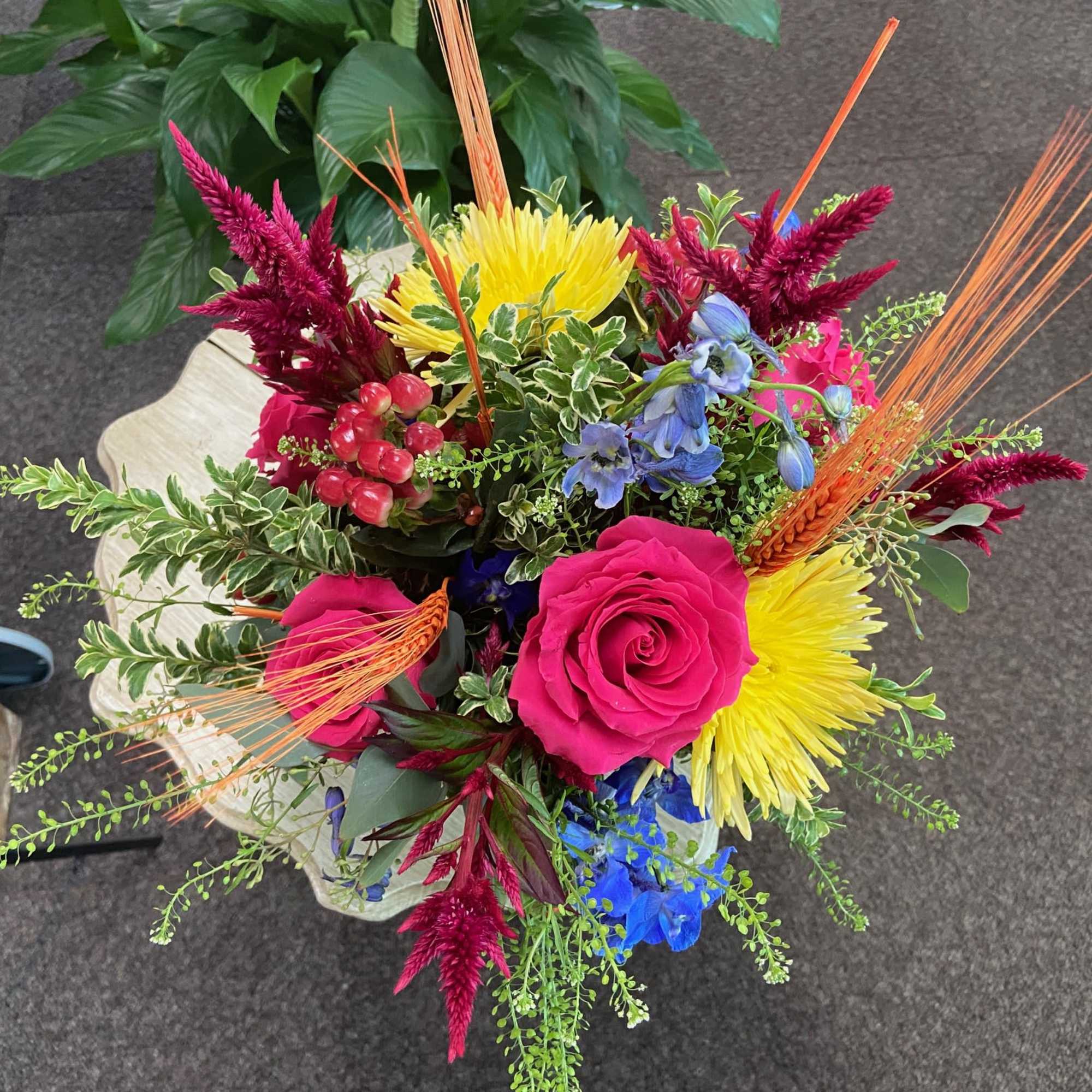 Bright mixed arrangement with hot pink roses, yellow mums, blue blooms, and red spikes in a light pedestal vase