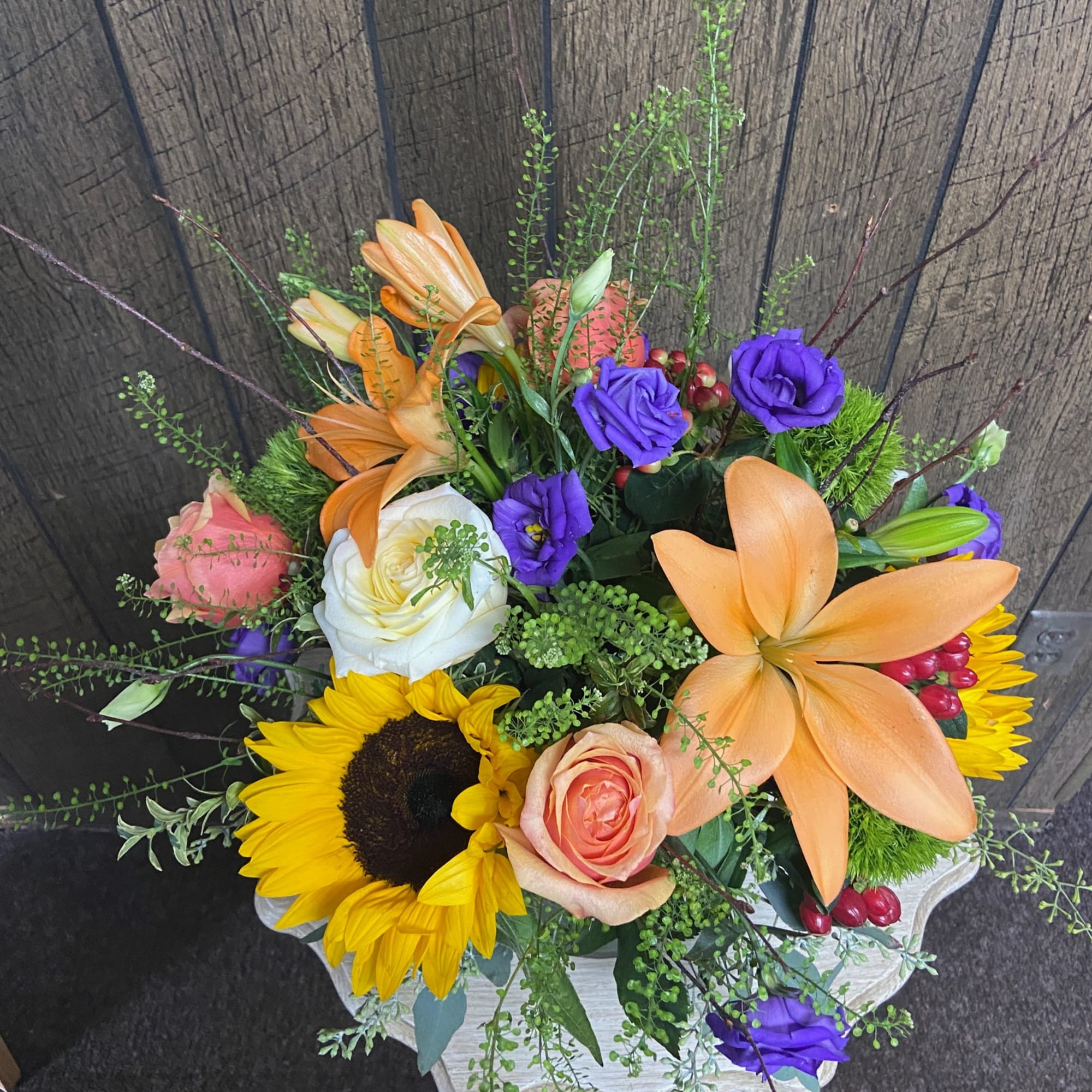 Mixed arrangement of orange lilies, sunflowers, roses, and purple lisianthus in a round container