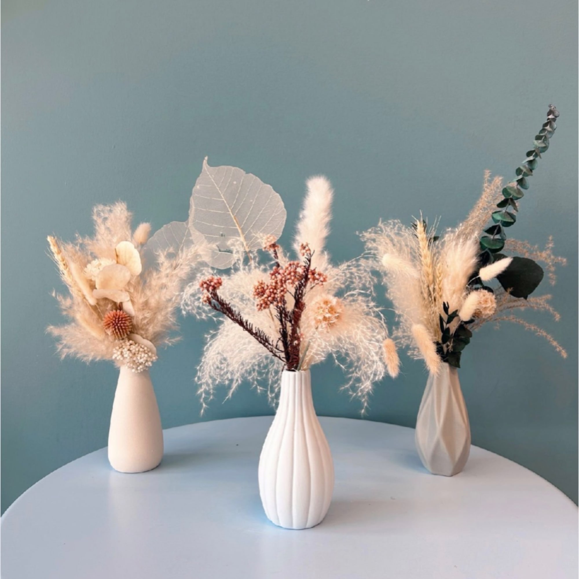 Three dried floral arrangements in white vases on a table