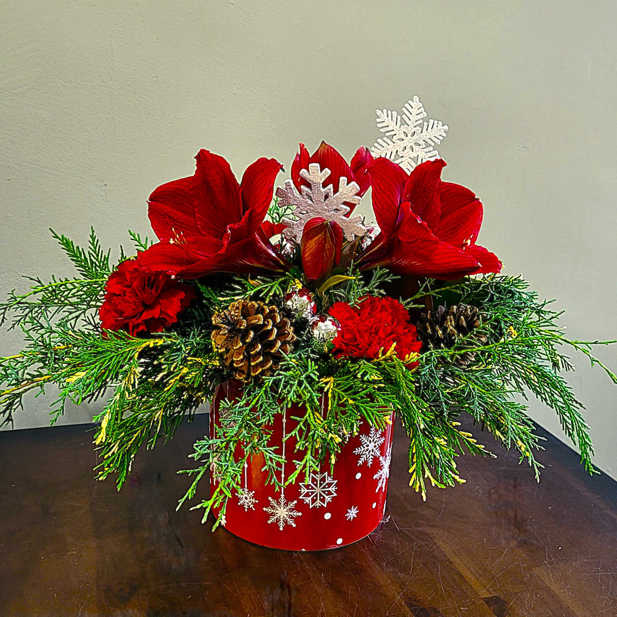 Low red holiday arrangement with red amaryllis, red carnations, evergreens, and pine cones in a snowflake container.