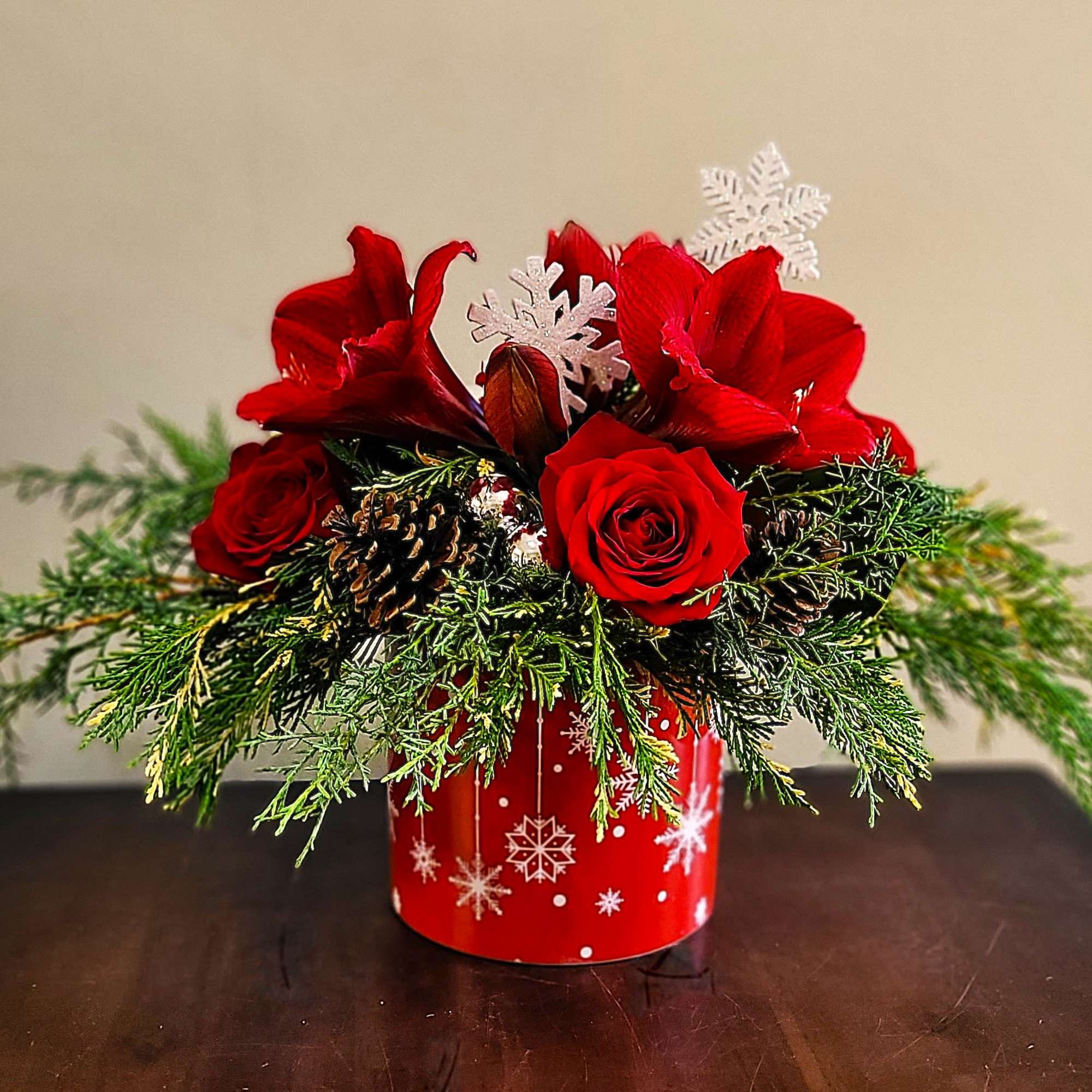 Low red rose and amaryllis arrangement with evergreens in a red snowflake container with pine cones and snowflake picks