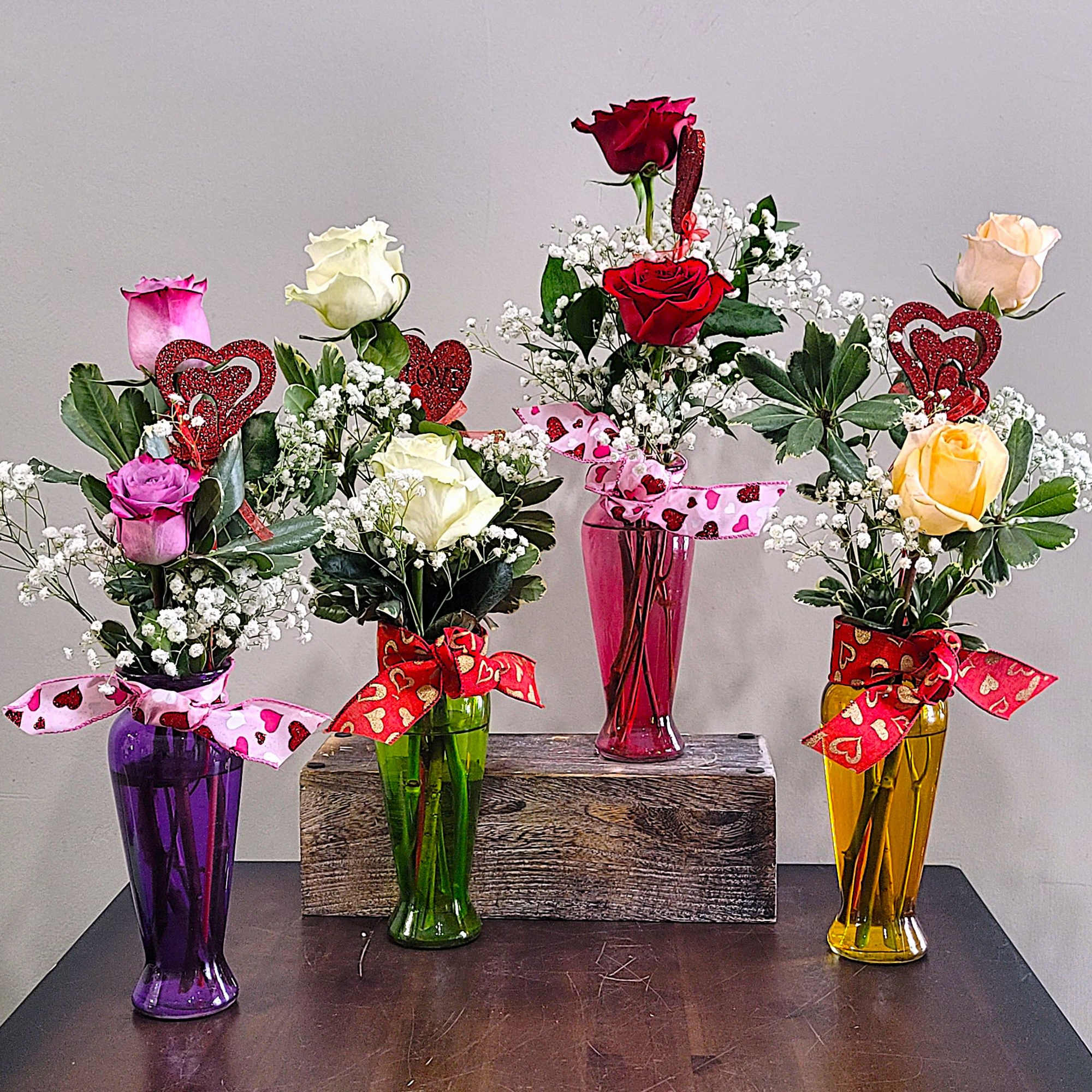 Four single-rose bud vase arrangements with baby's breath, heart picks, and ribbons in colored glass vases