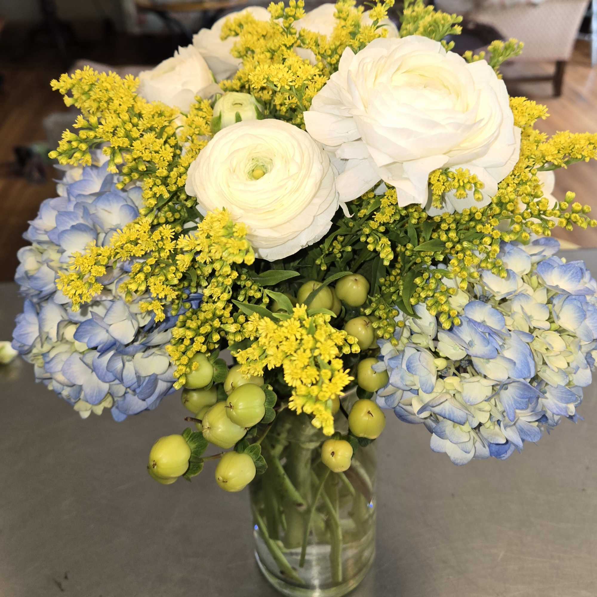 Bouquet of white ranunculus, blue hydrangea, and yellow filler in a glass vase
