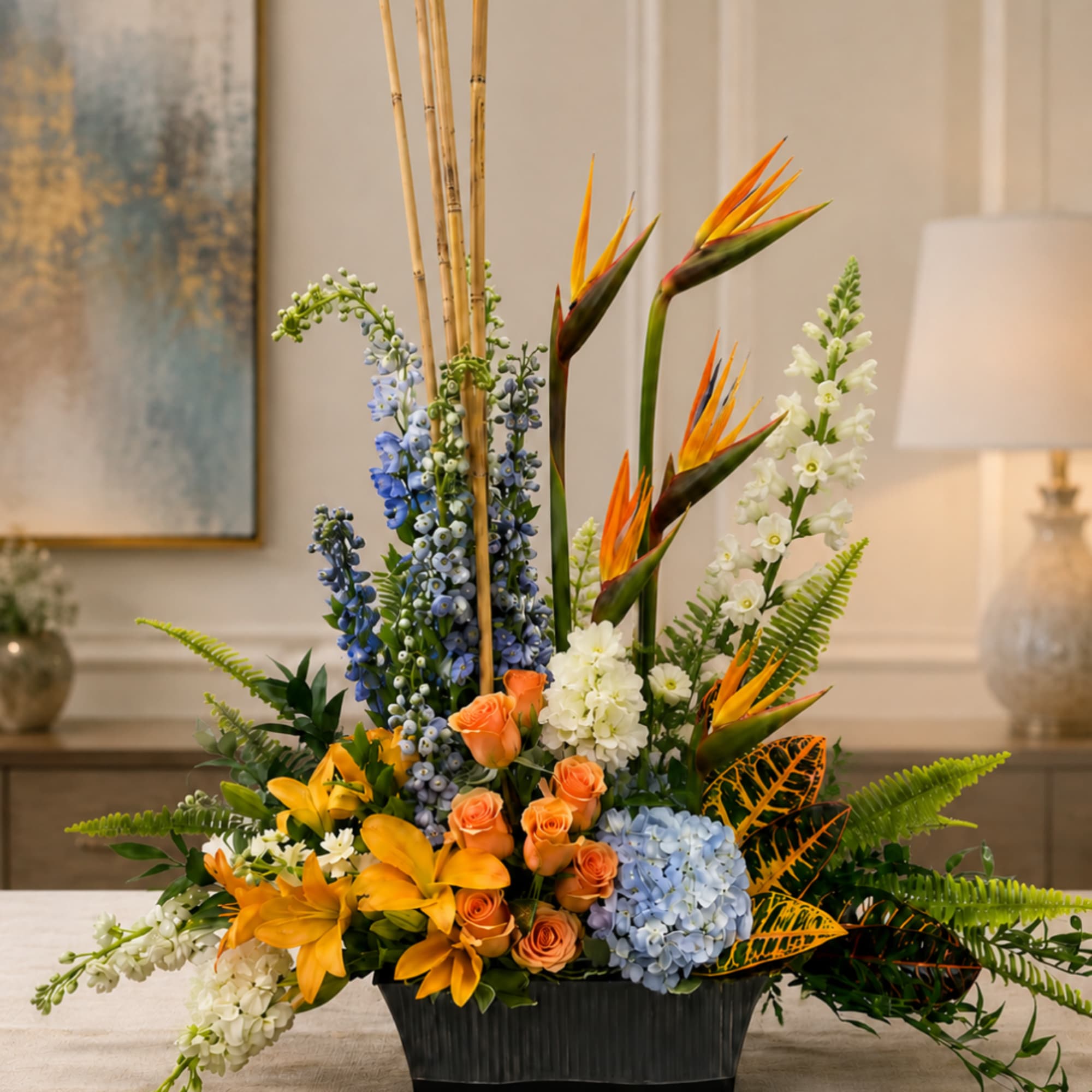 Tropical floral arrangement with orange roses, blue hydrangeas, and bird of paradise in a black container