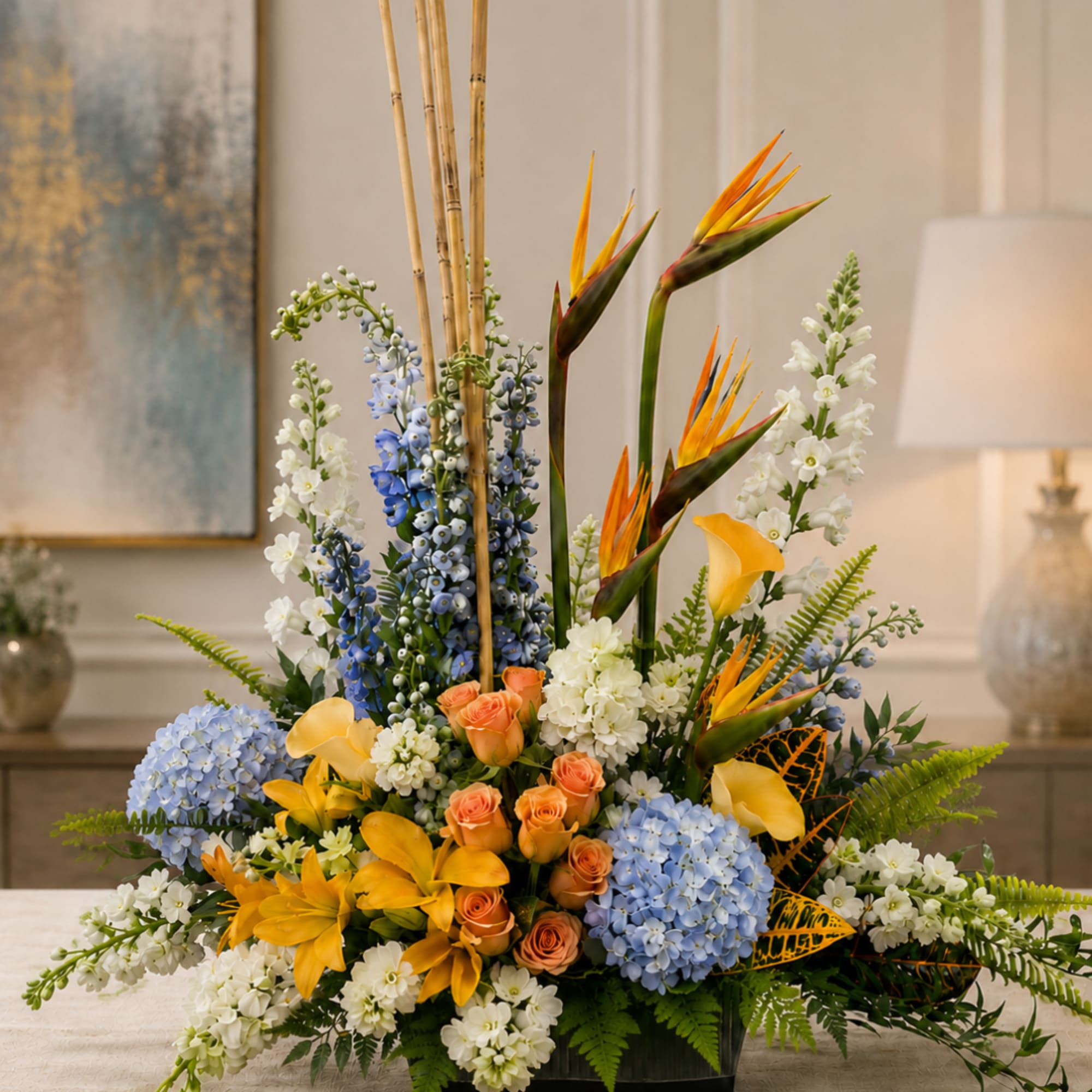Tropical floral arrangement with blue hydrangeas, orange birds of paradise, and peach roses in a low container.