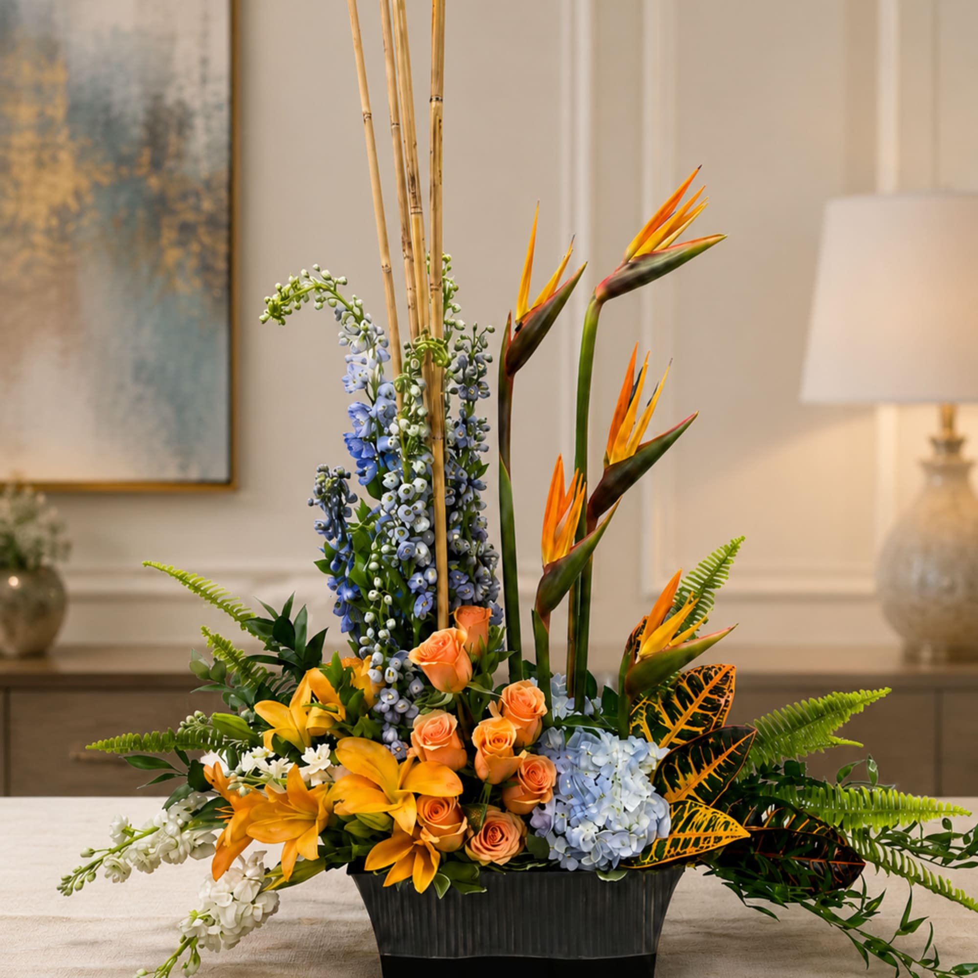 Tropical floral arrangement with orange birds of paradise, roses, lilies, and blue blooms in a black vase