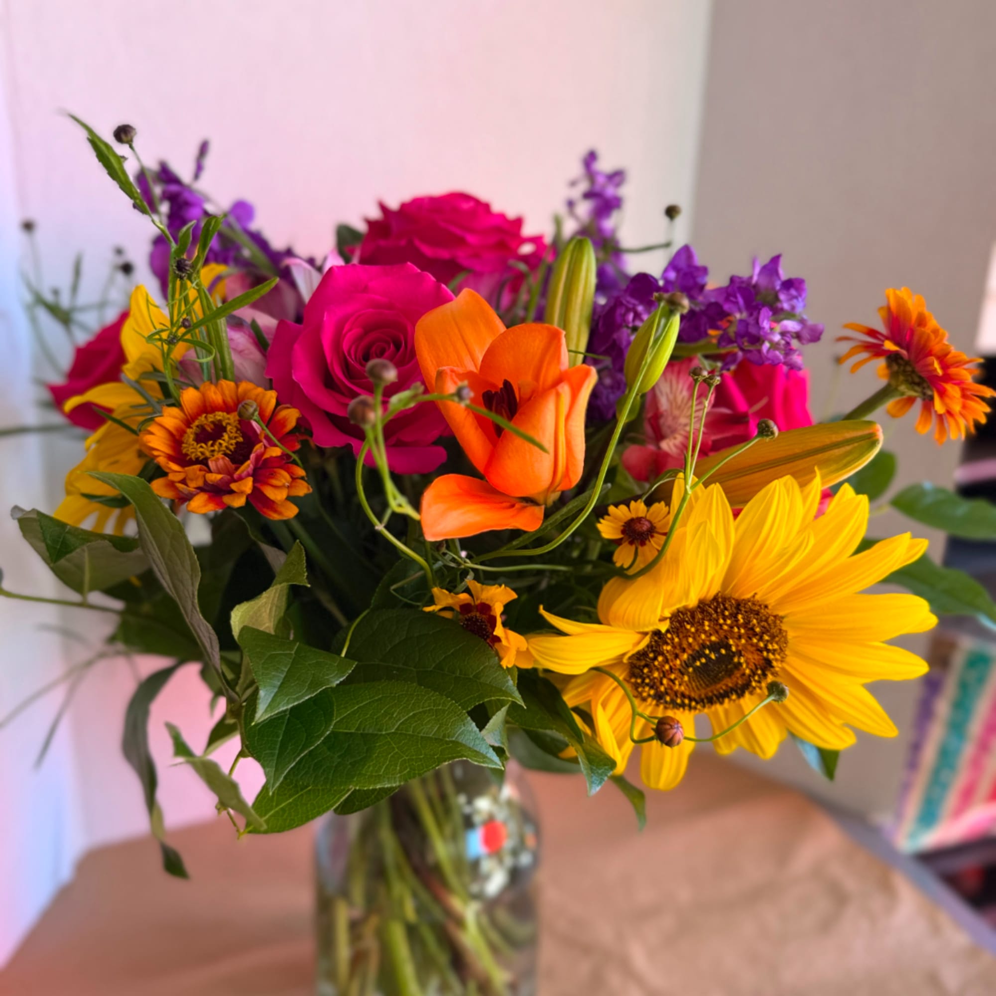 Mixed bouquet with roses, lilies, sunflowers, and purple blooms in a glass vase