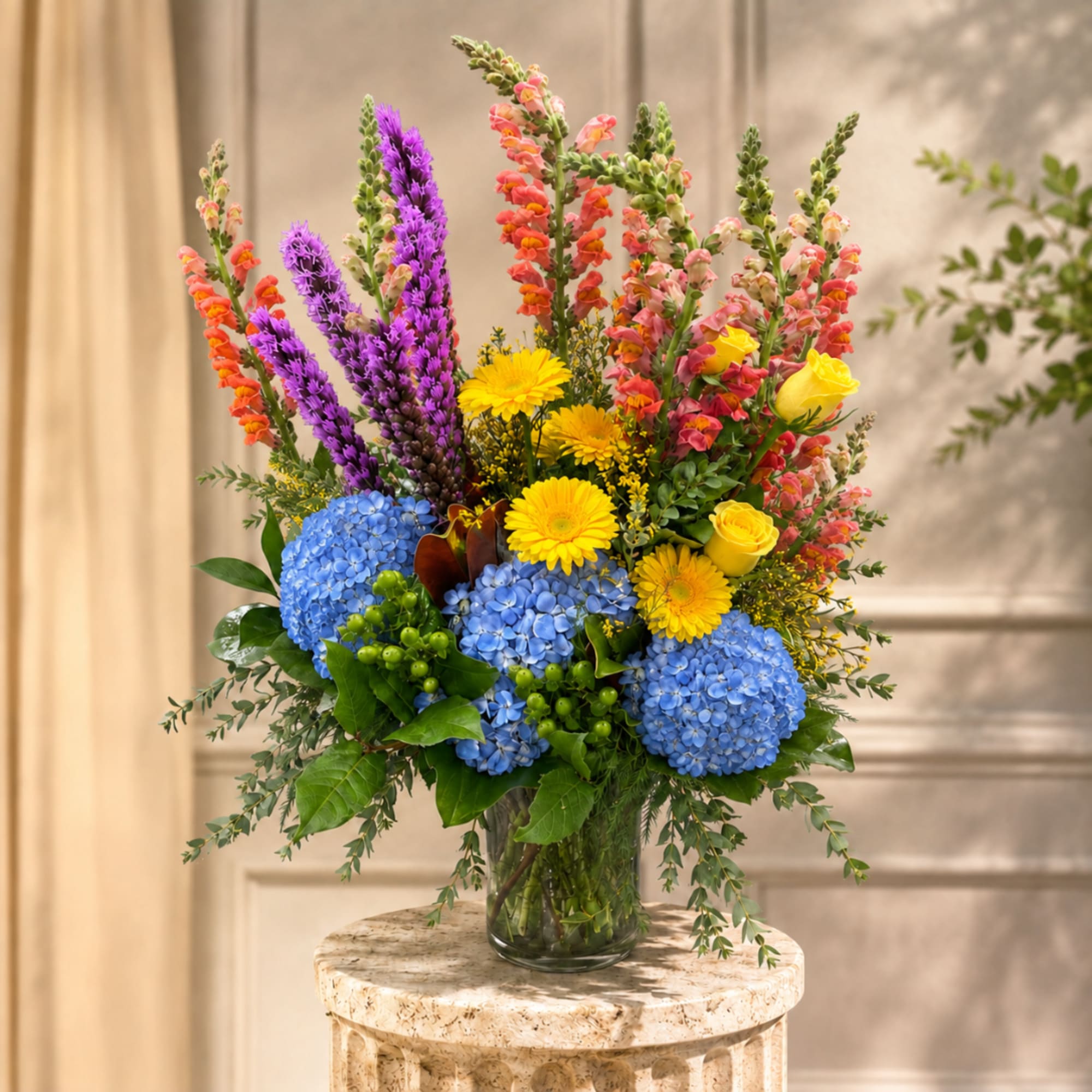 Tall mixed bouquet in a clear glass vase with blue hydrangeas, yellow daisies, and pink snapdragons