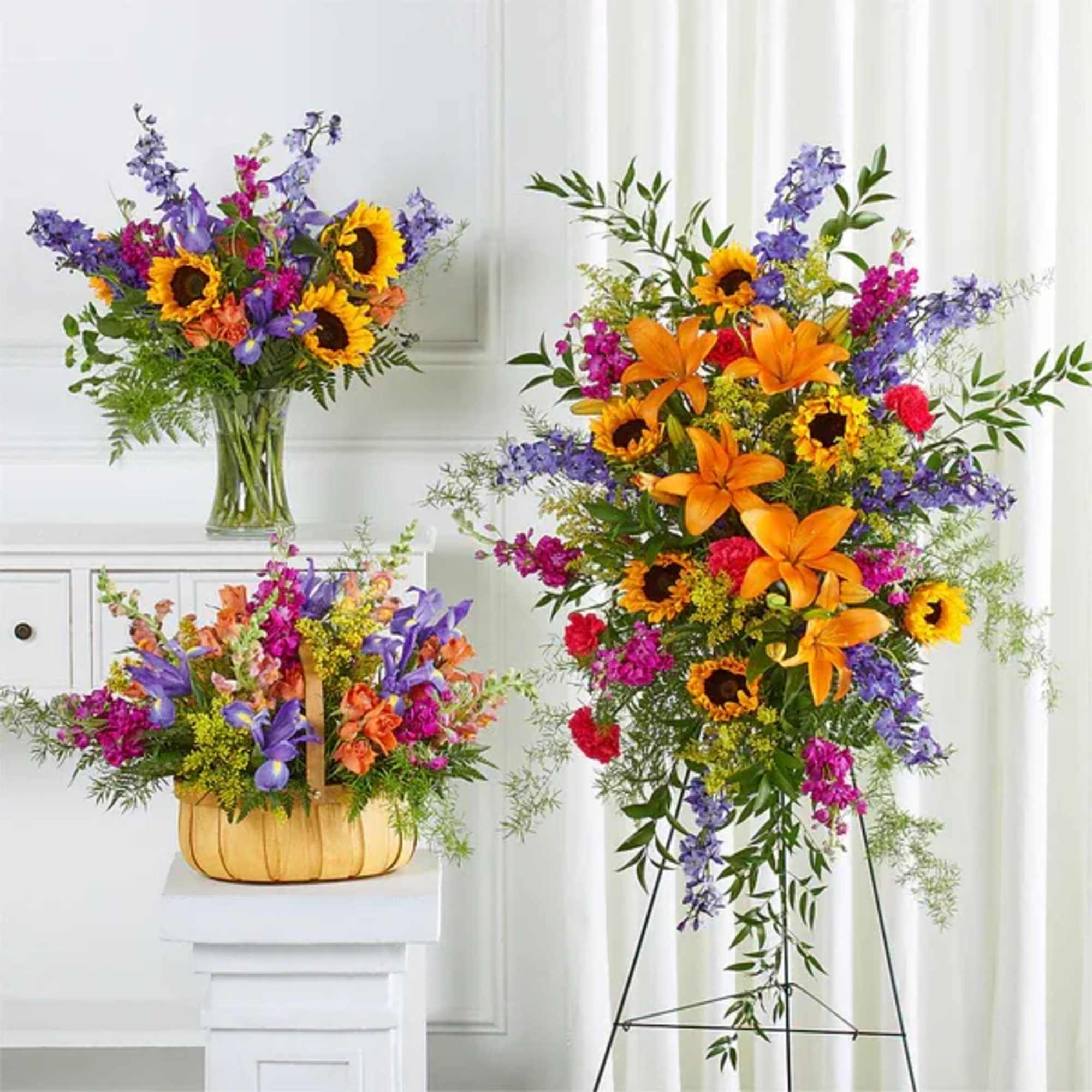 Three bright mixed floral arrangements with sunflowers and lilies in a vase, basket, and standing easel spray.