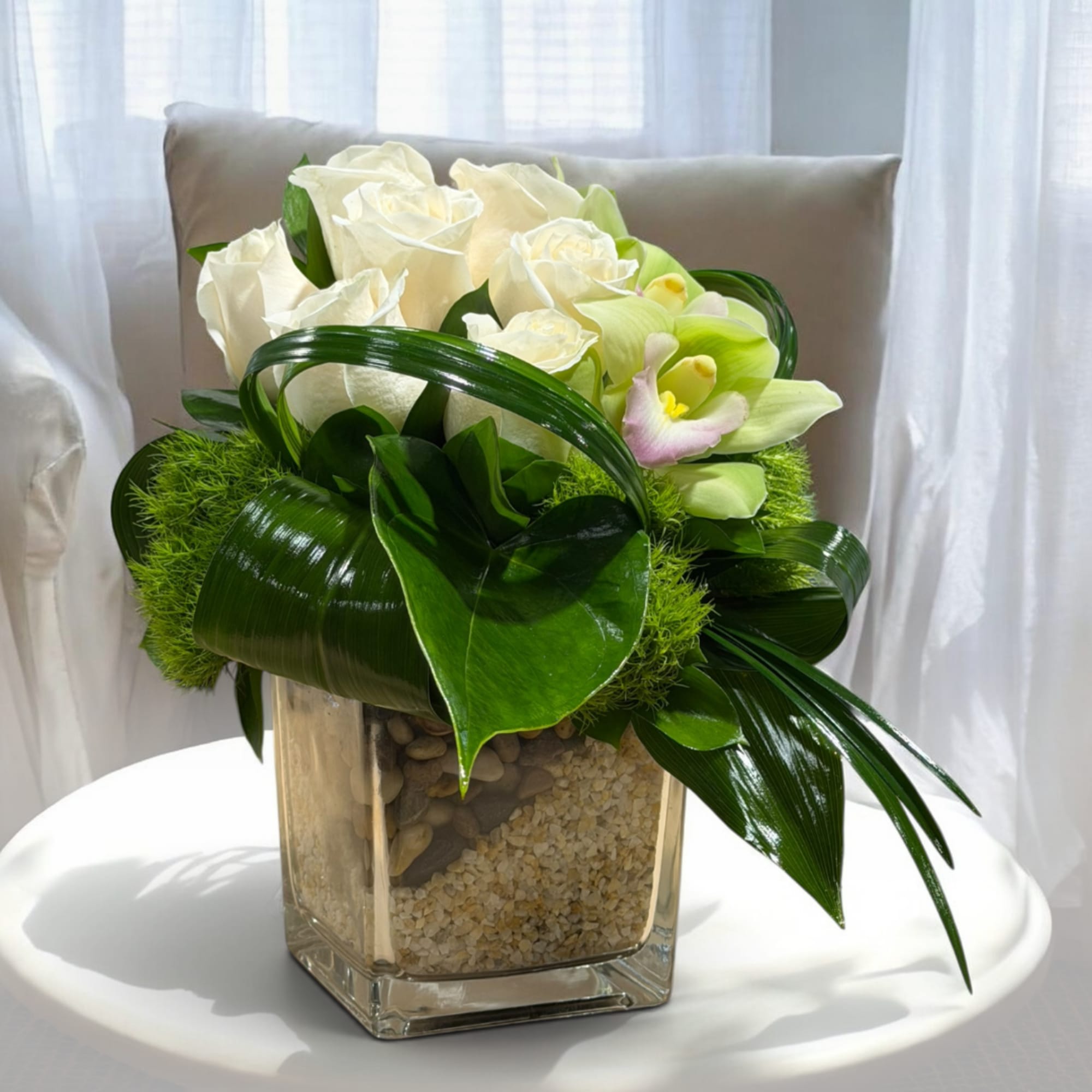 White roses and pale green orchids in a square glass vase with decorative stones.