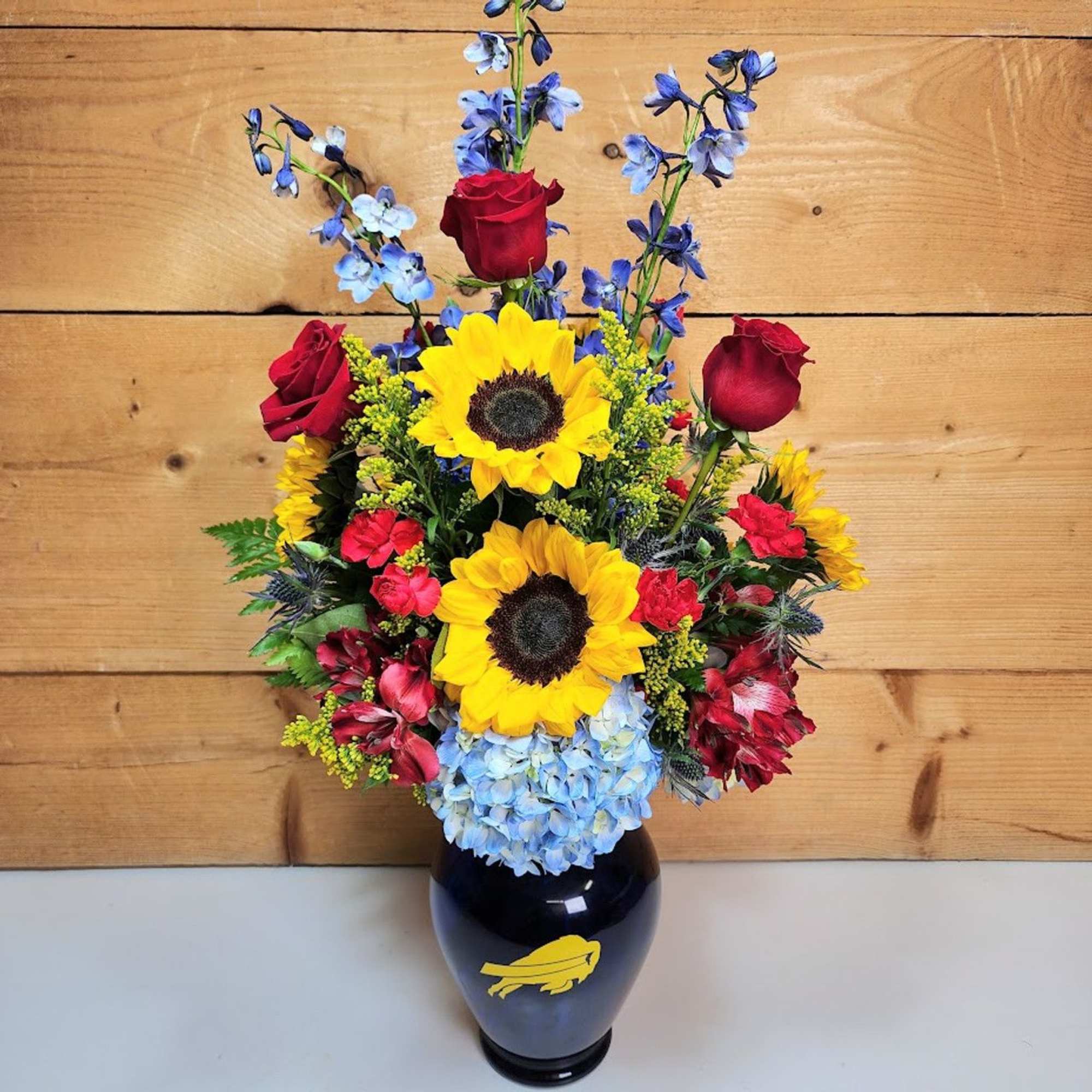 Mixed bouquet with sunflowers, red roses, blue flowers, and hydrangea in a dark vase