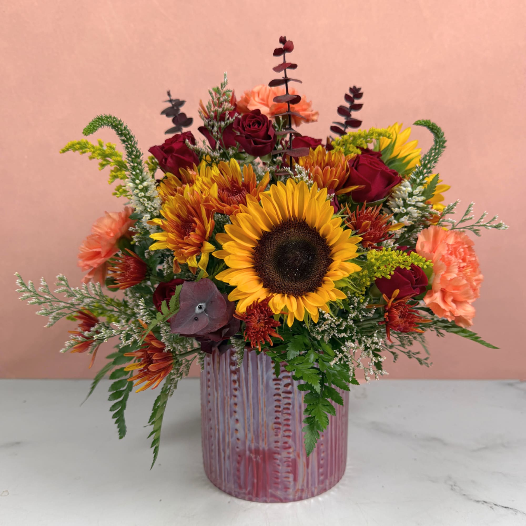 Mixed bouquet with sunflowers, red roses, and peach carnations in a pink vase