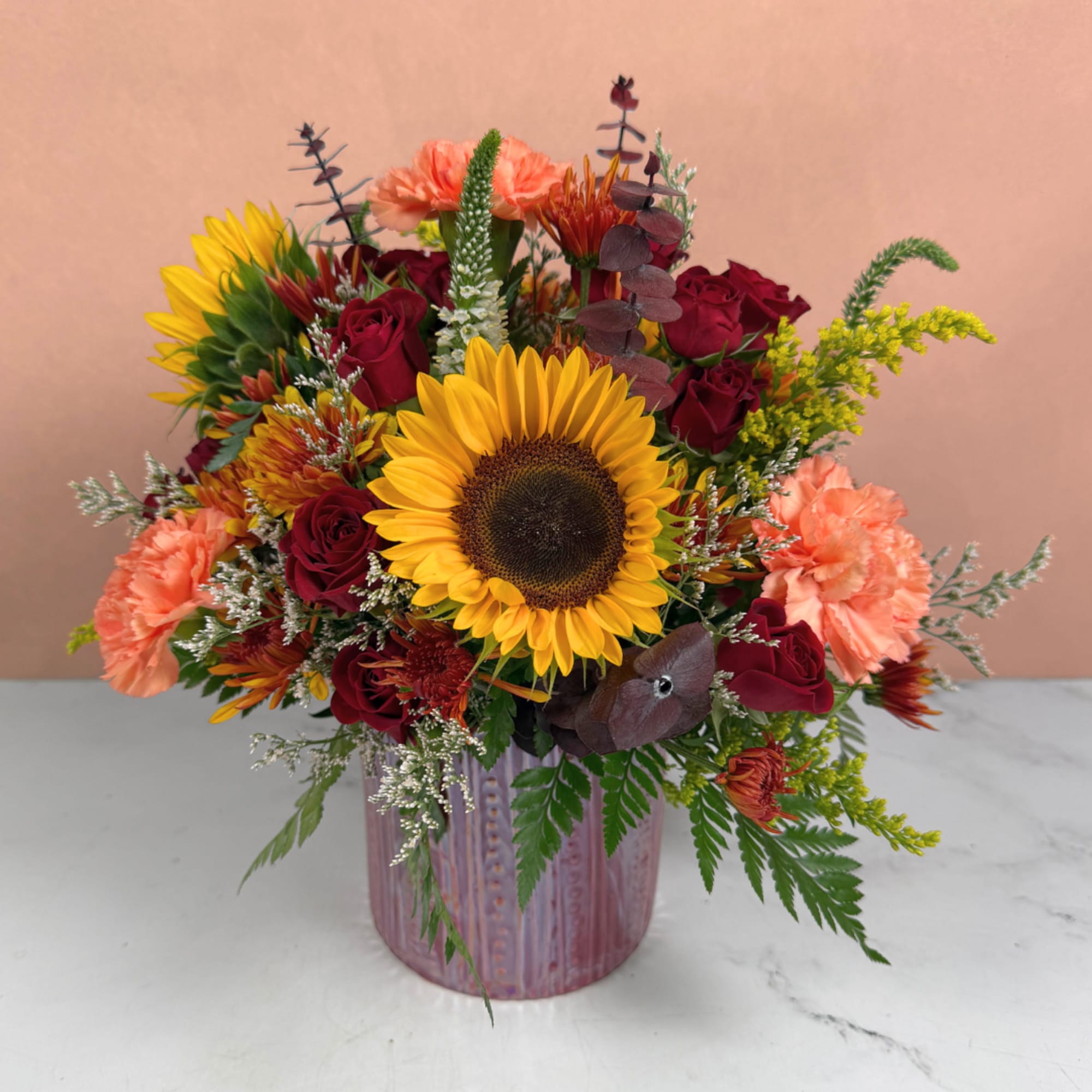 Mixed bouquet with a sunflower, red roses, and peach carnations in a pink vase