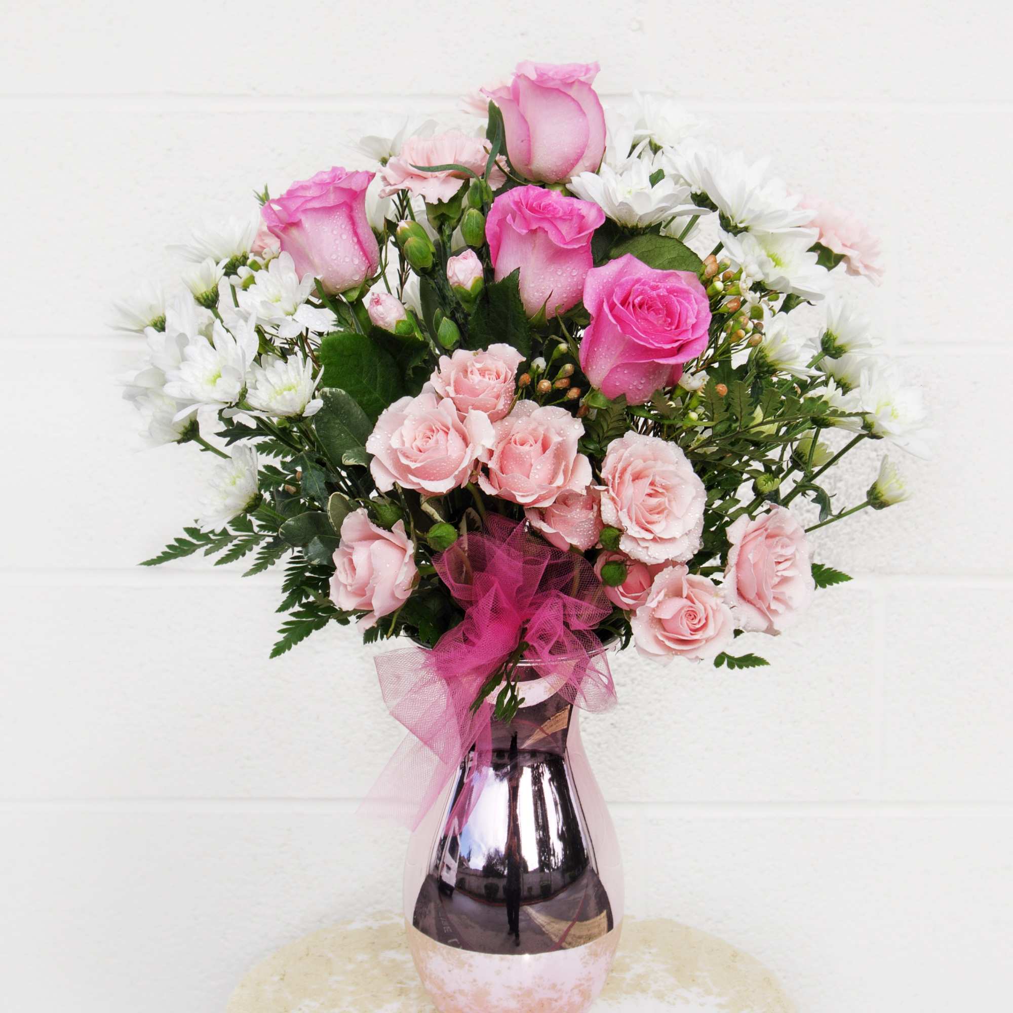 Pink roses and white daisies arranged in a metallic vase with a pink ribbon