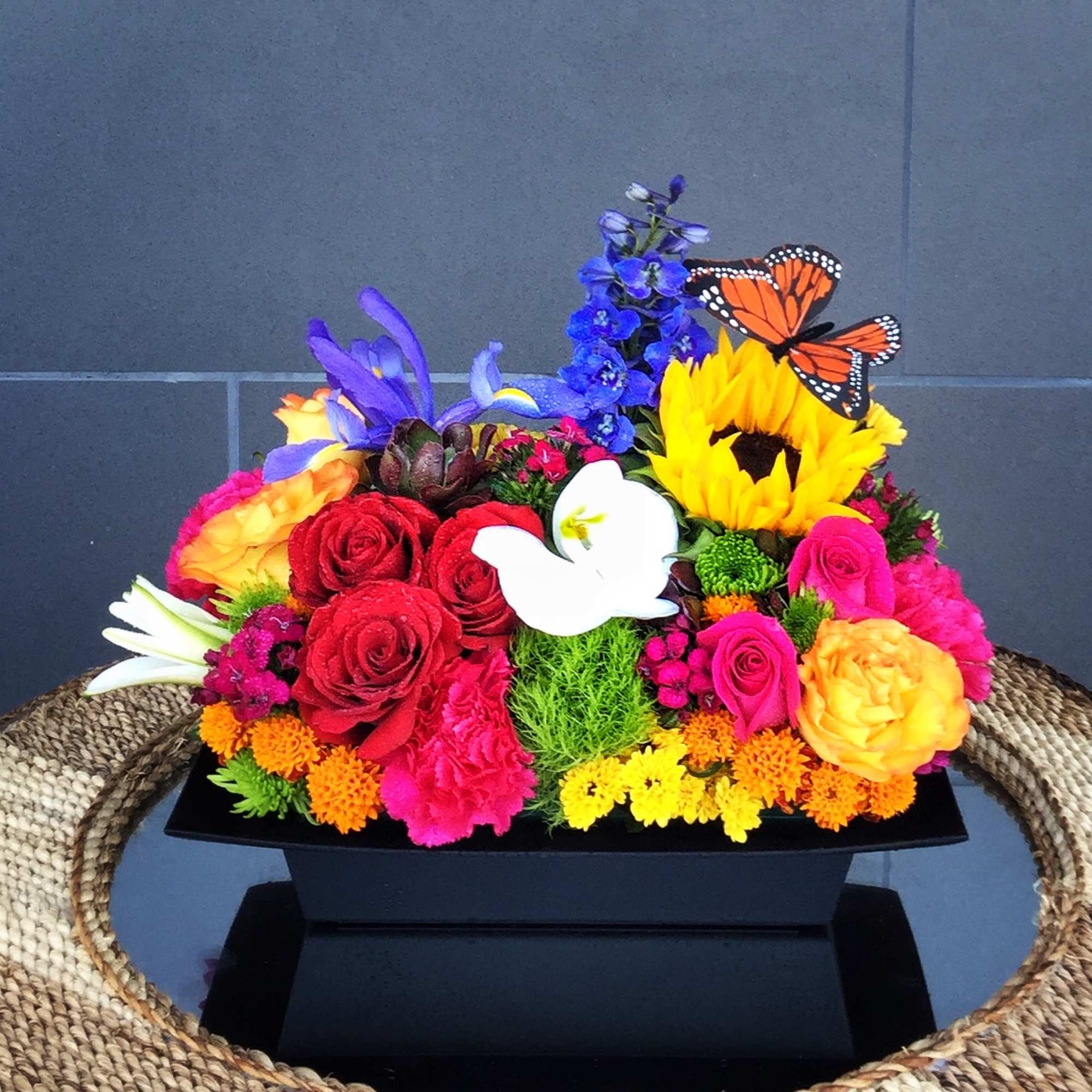 Colorful floral arrangement with roses, sunflowers, and butterflies in a black container