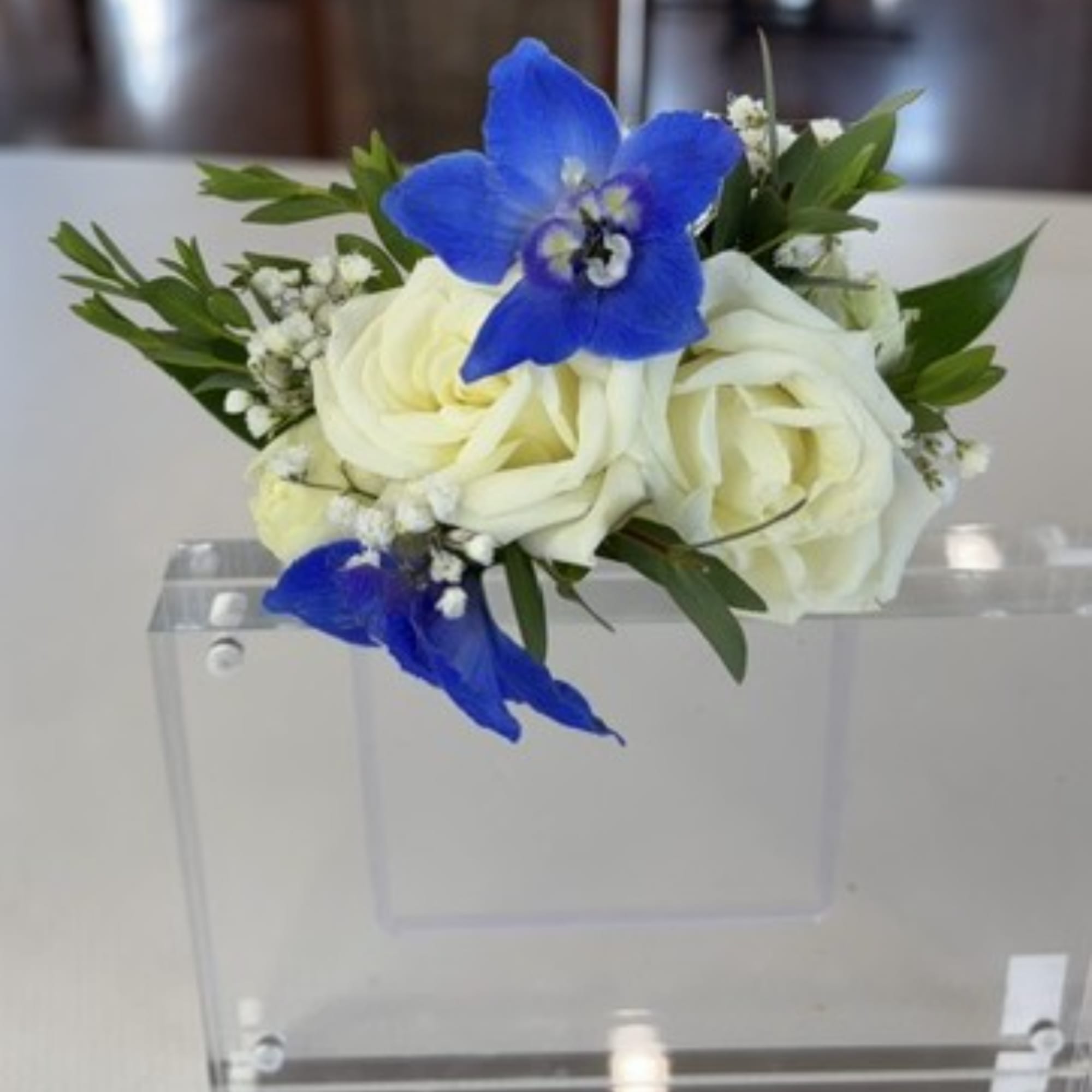 White roses with blue orchids and greenery in a clear acrylic holder