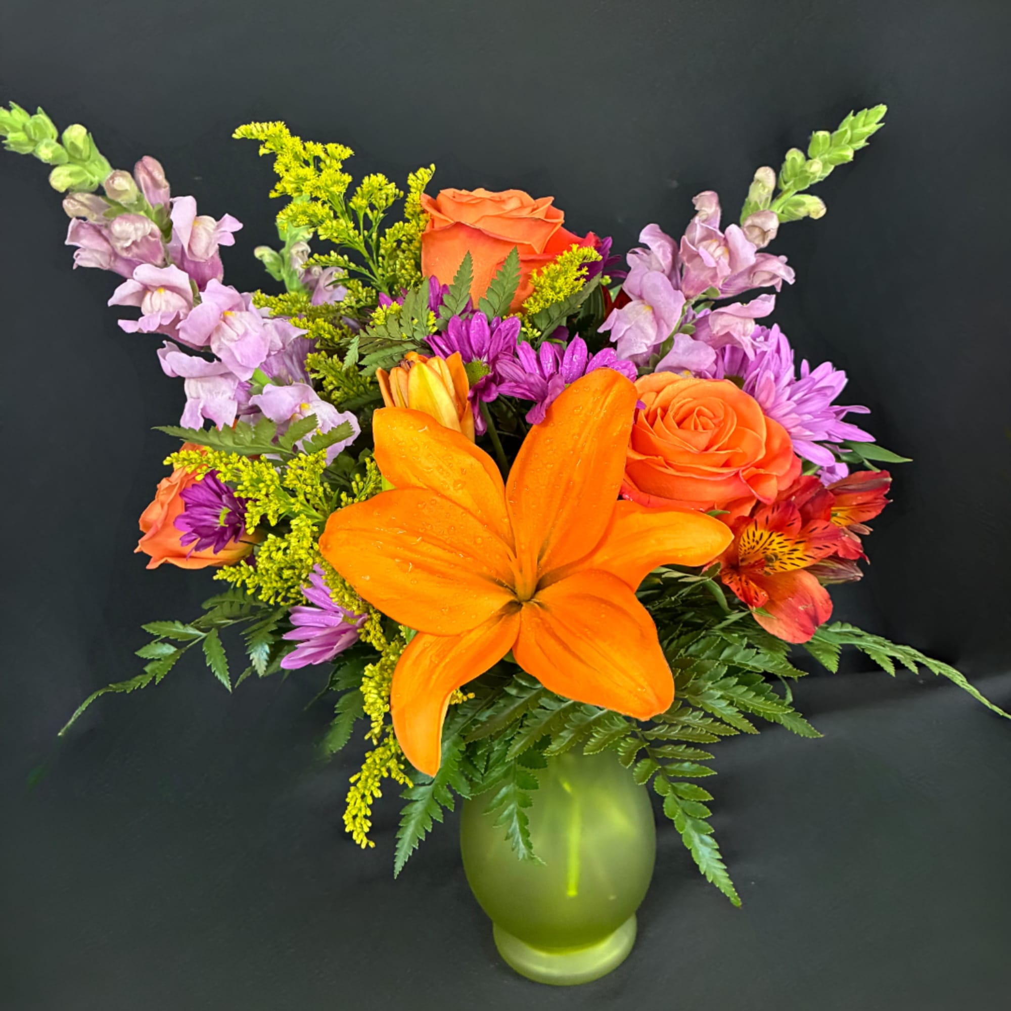Orange lilies and roses in a green vase with purple flowers