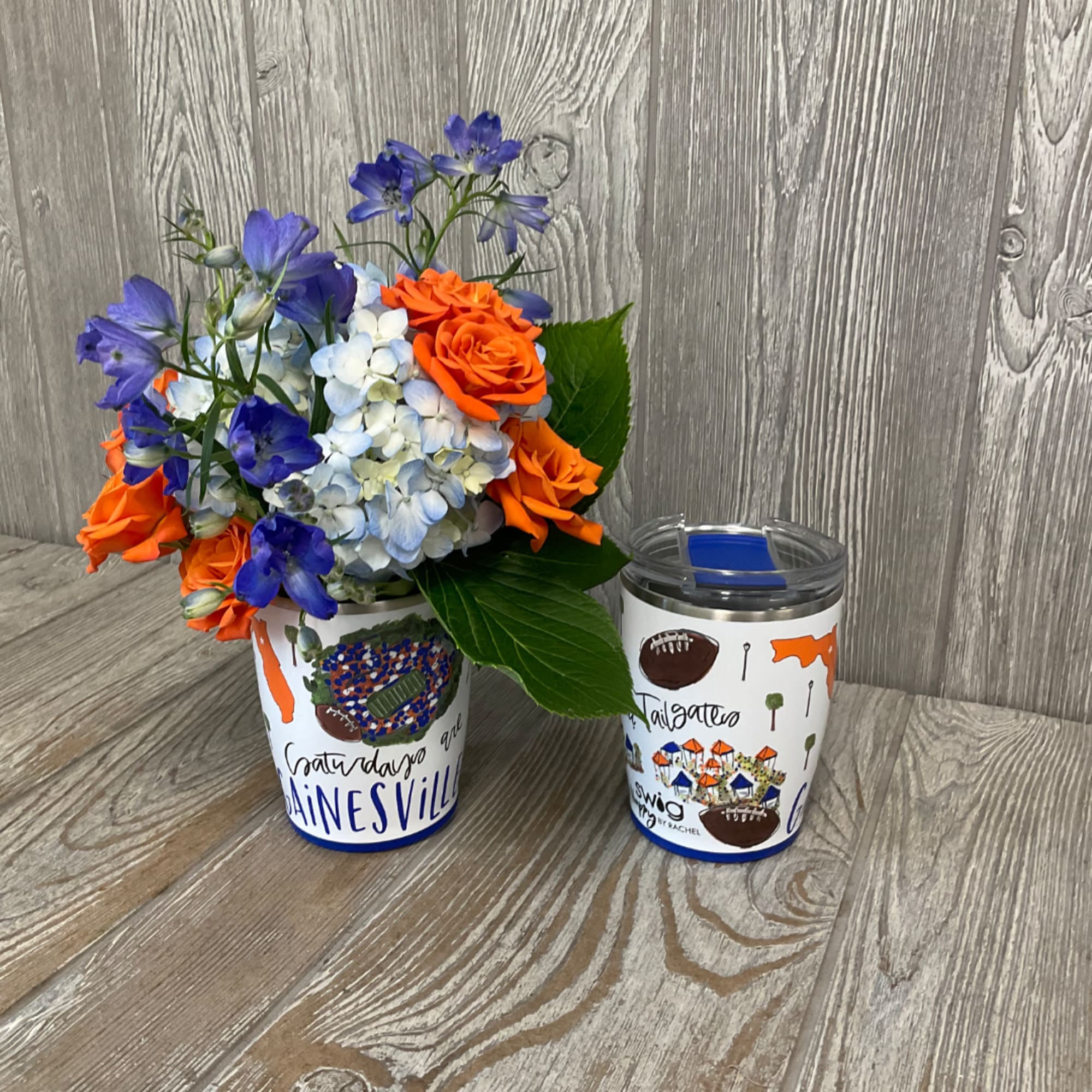 Orange roses and blue flowers in a decorated cup beside a matching tumbler