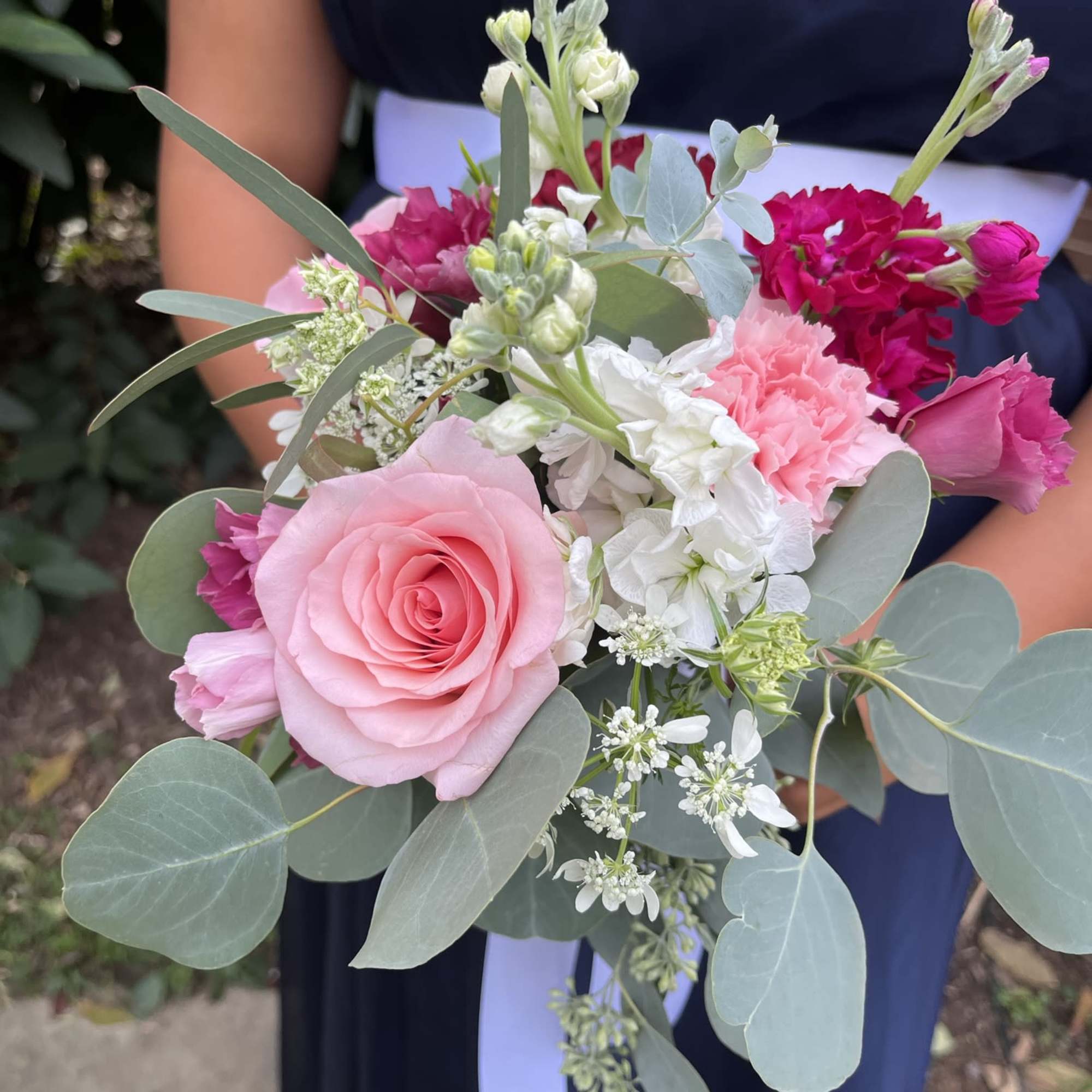 Hand-tied bouquet of pink roses, carnations, and white blooms with eucalyptus, held against a navy dress.