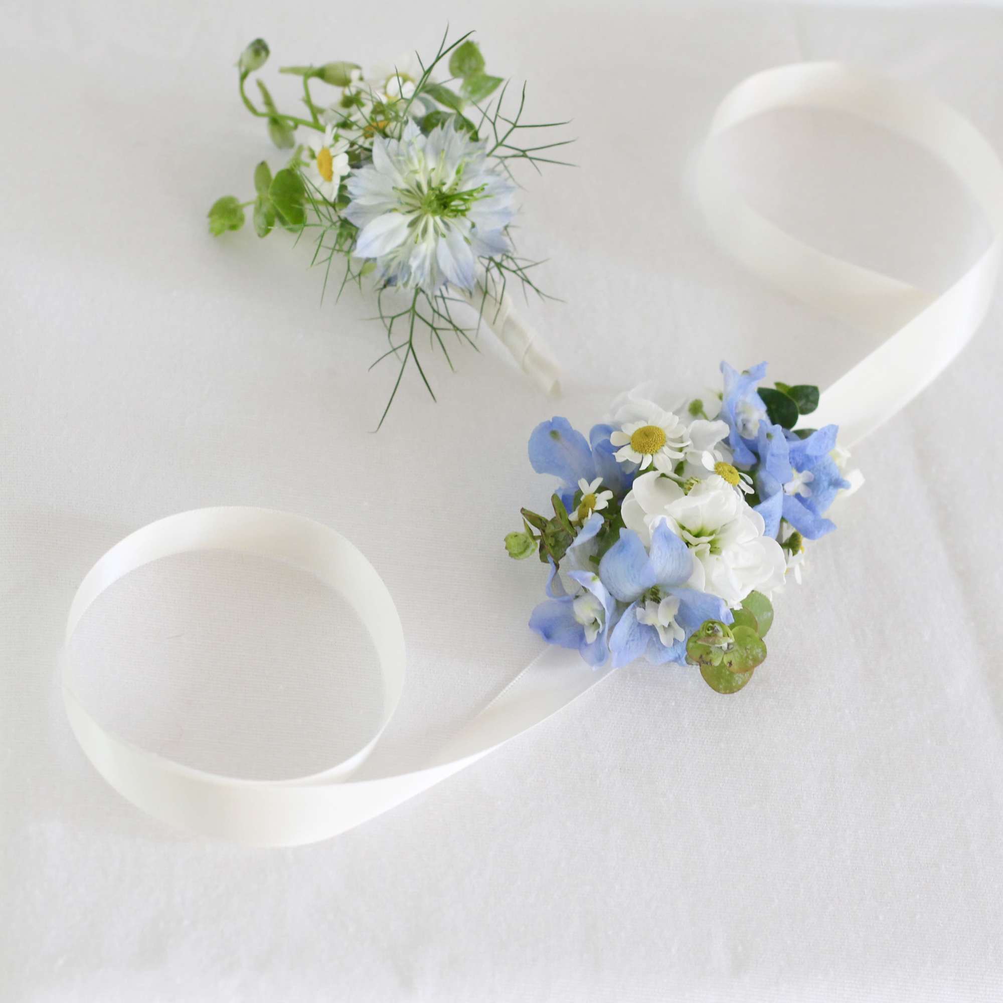 Two small floral boutonnieres with blue and white flowers on white ribbon