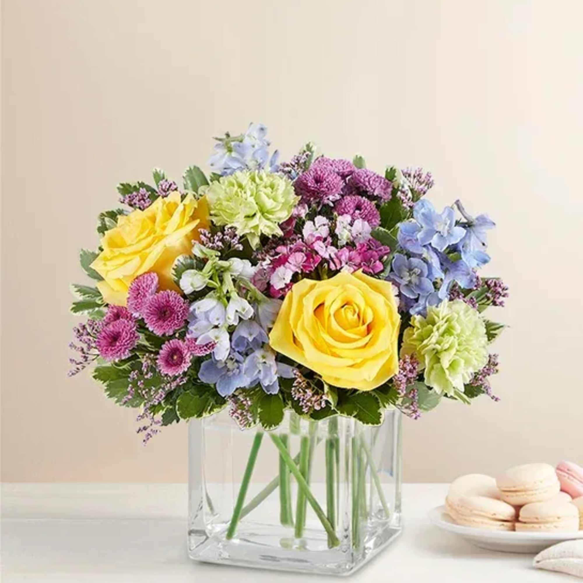 Bouquet of yellow roses, blue flowers, and pink blooms in a clear square vase