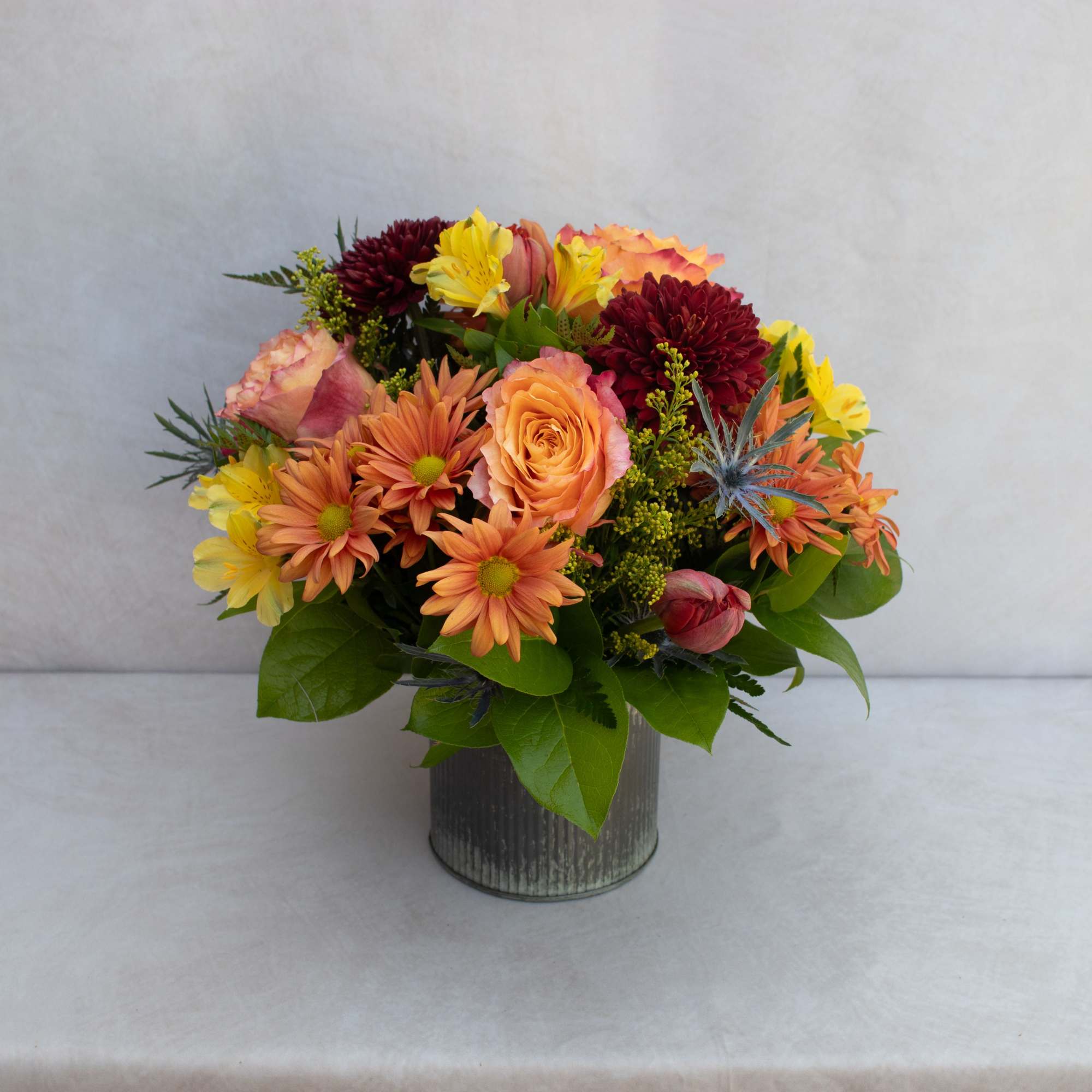 Mixed bouquet of orange, yellow, and burgundy flowers in a metal vase