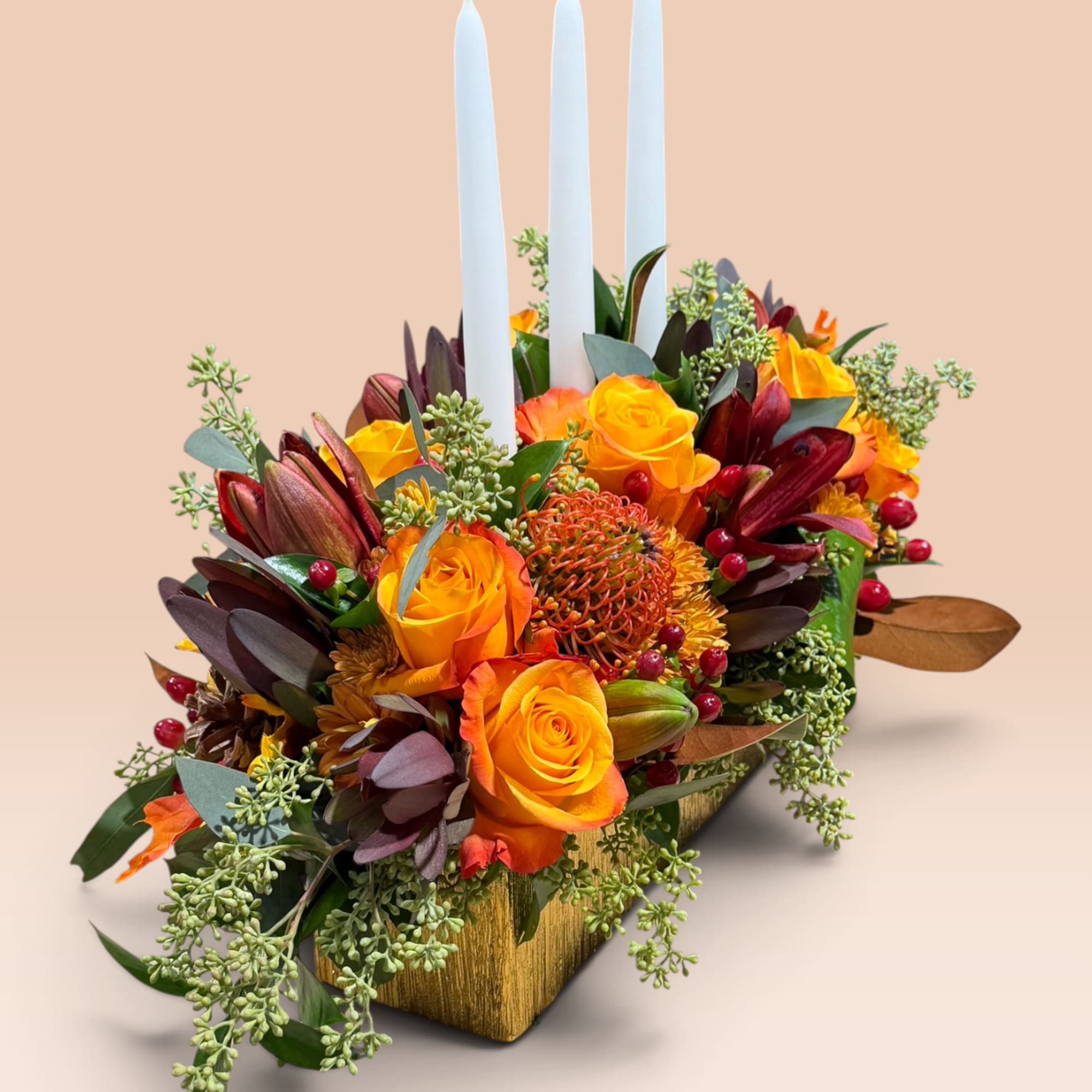 Orange roses and mixed flowers arranged with three white candles in a gold container