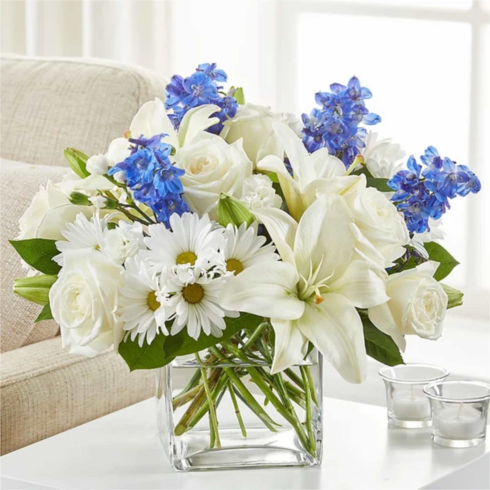White roses, lilies, and daisies with blue flowers in a clear glass vase