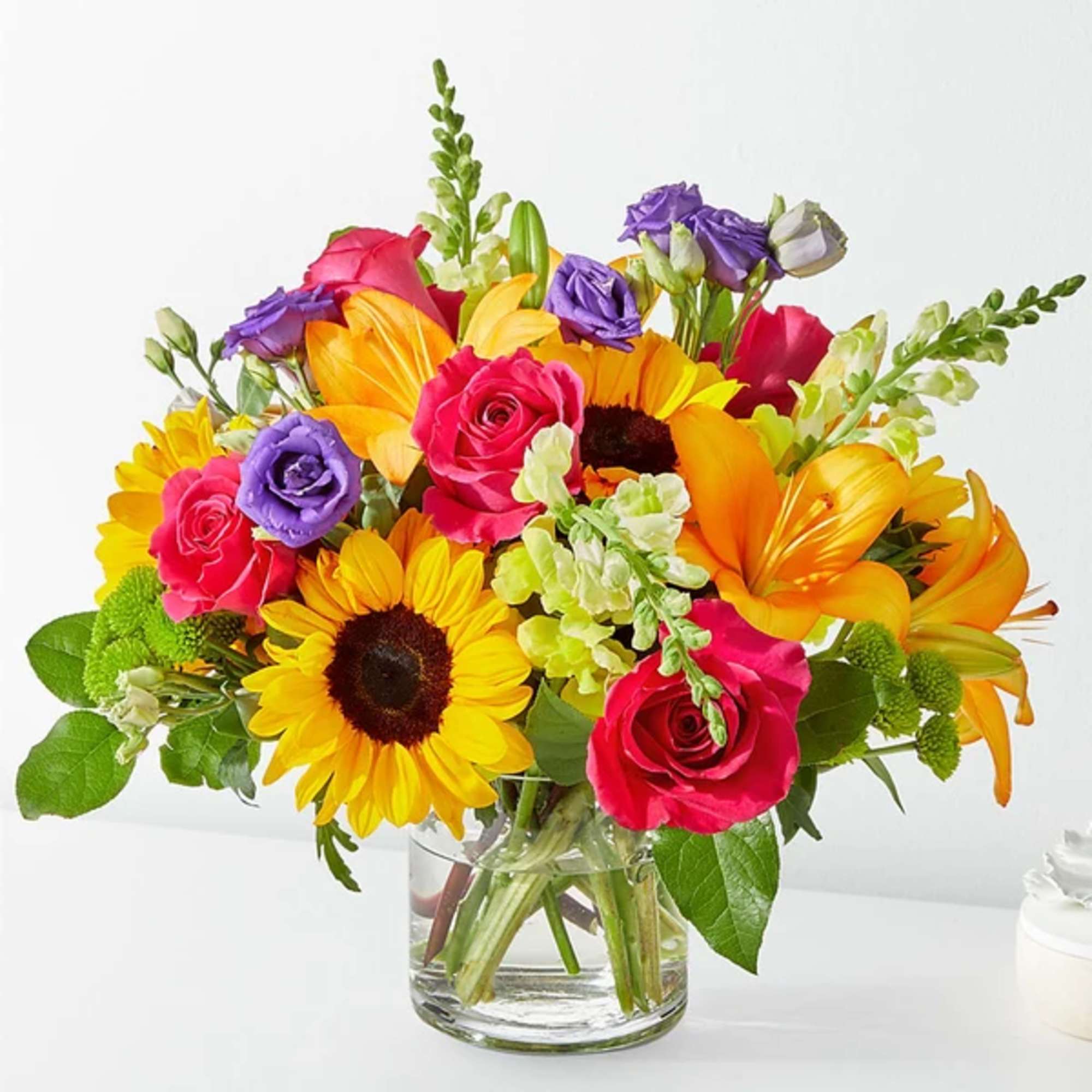 Mixed bouquet of sunflowers, roses, lilies, and purple blooms in a glass vase